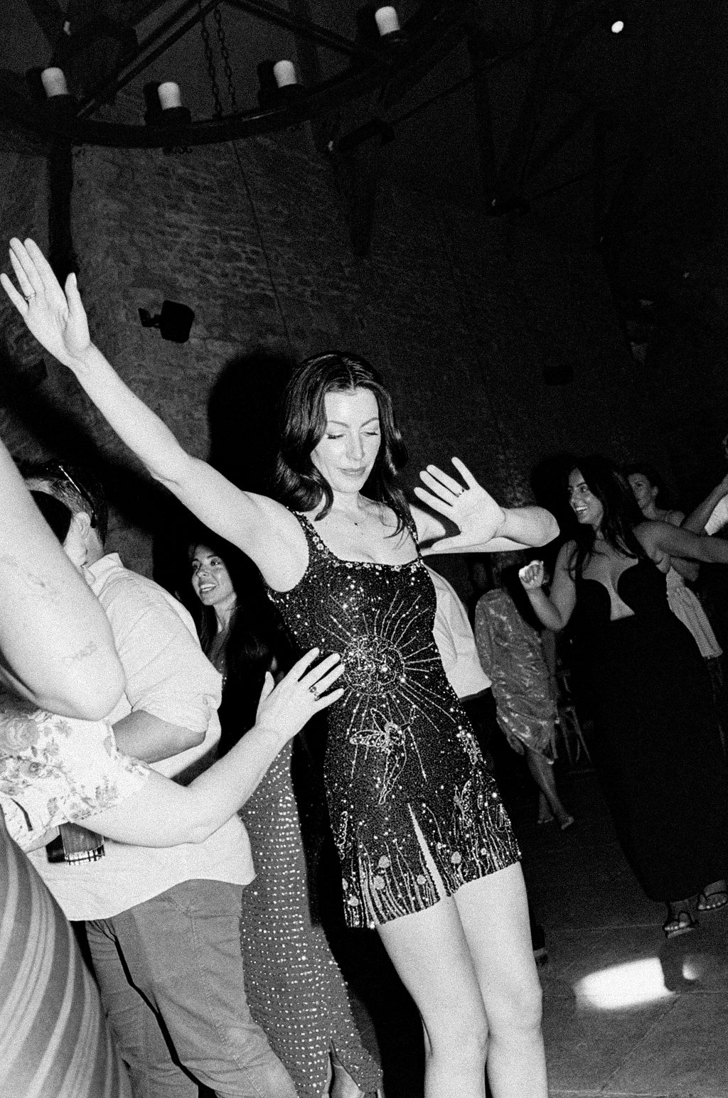 Woman in sequined mini dress dancing with arms raised at a lively party in a dimly lit venue.