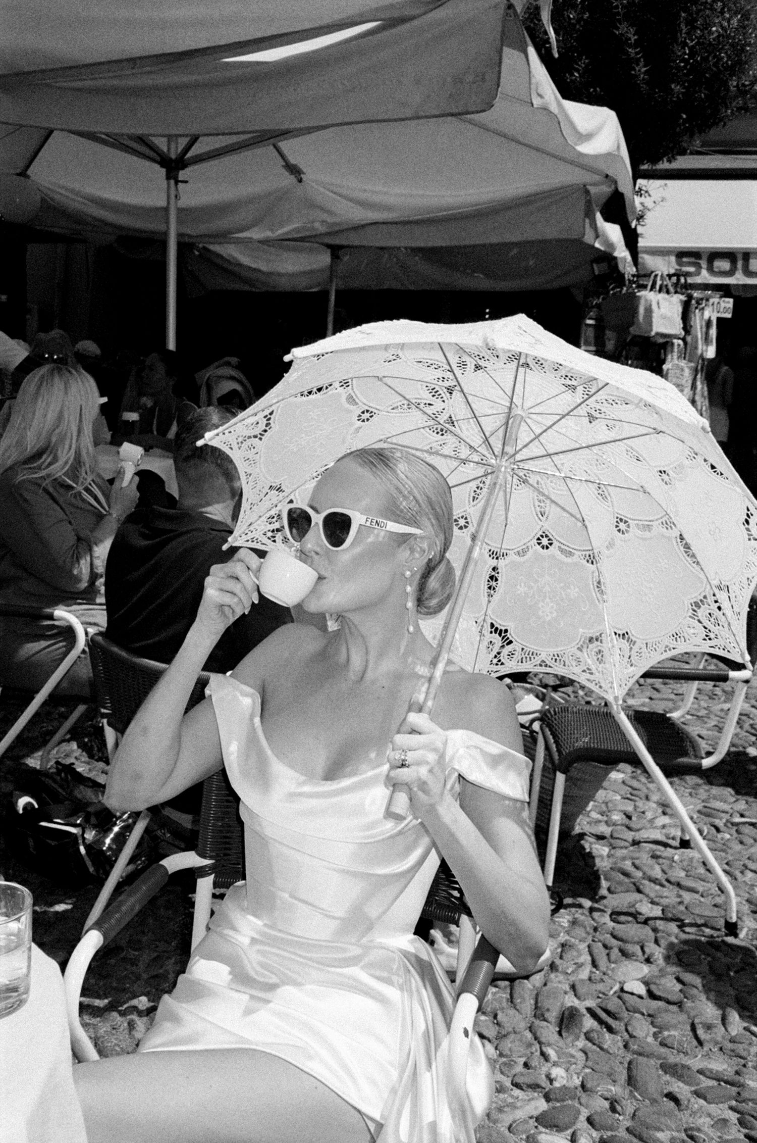 Elegant woman in white dress sipping coffee under lace parasol at outdoor cafe, black and white photo.