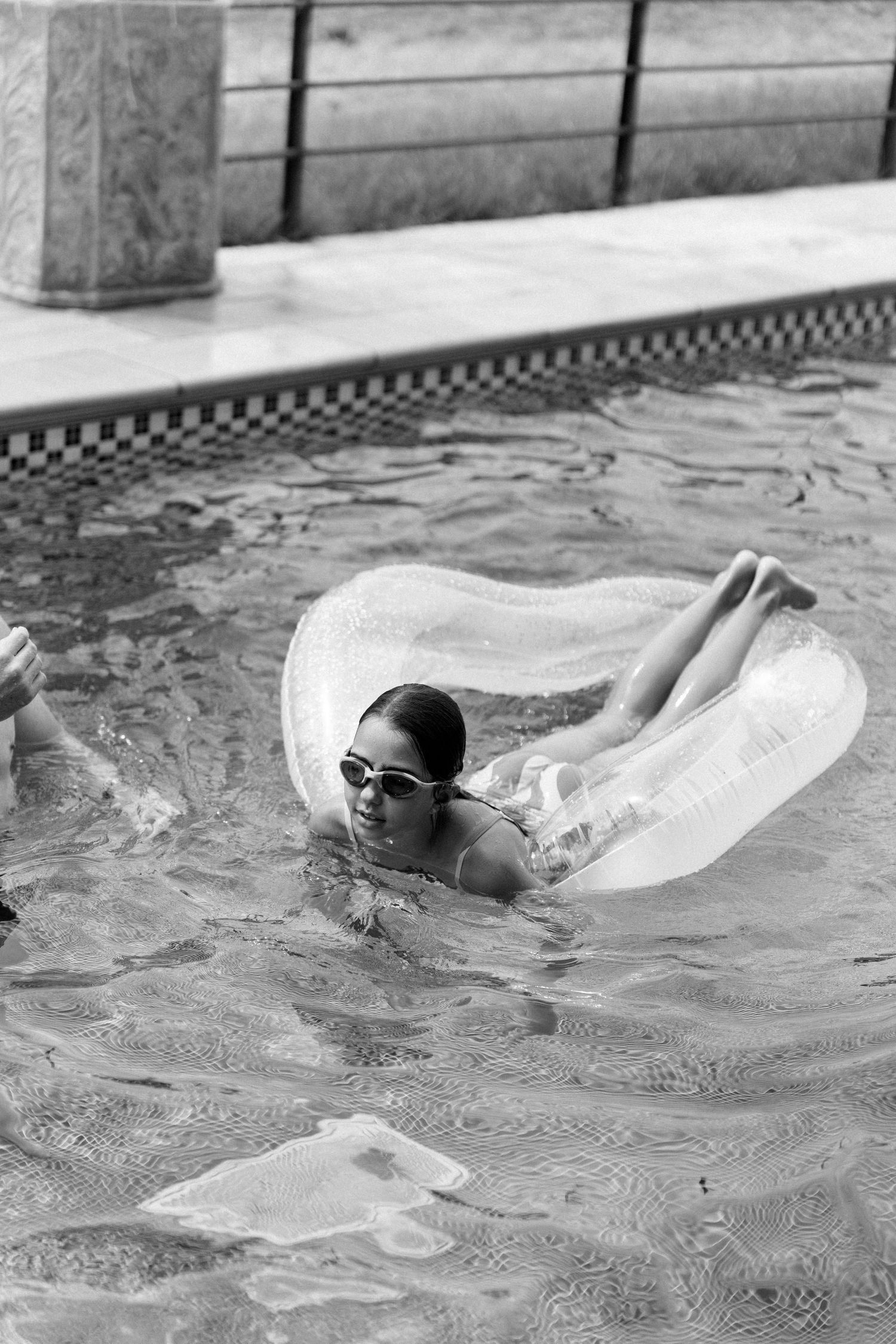 Black and white photo of woman floating in clear inflatable ring in swimming pool wearing sunglasses.