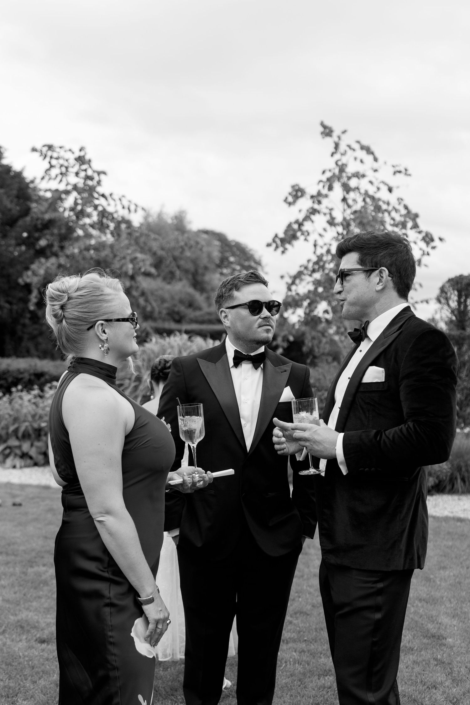 Three elegantly dressed wedding guests in black tie attire conversing and holding champagne glasses outdoors.