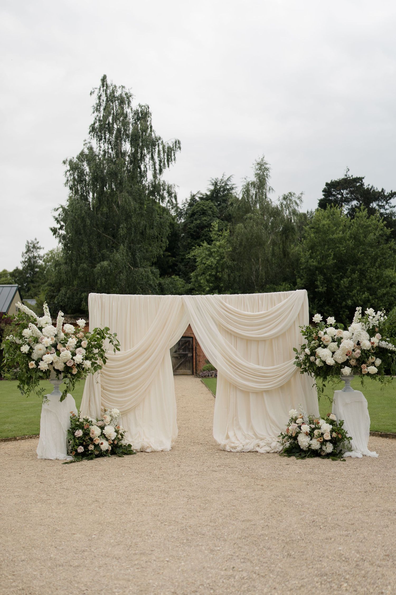 Elegant outdoor wedding ceremony arch draped in white fabric with floral arrangements on a gravel path.
