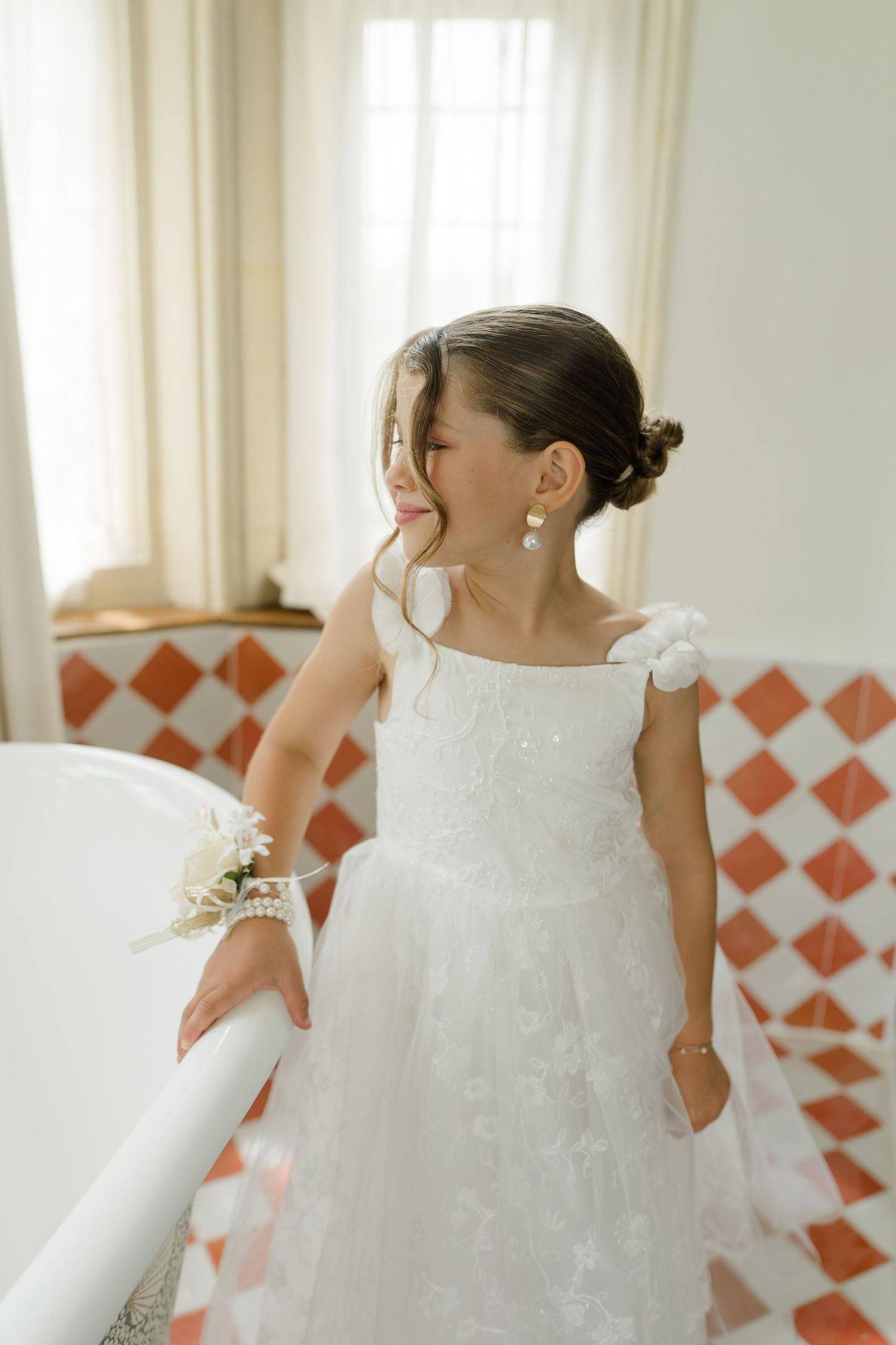 Young flower girl in white lace dress with floral earrings standing by a bathtub in tiled bathroom.