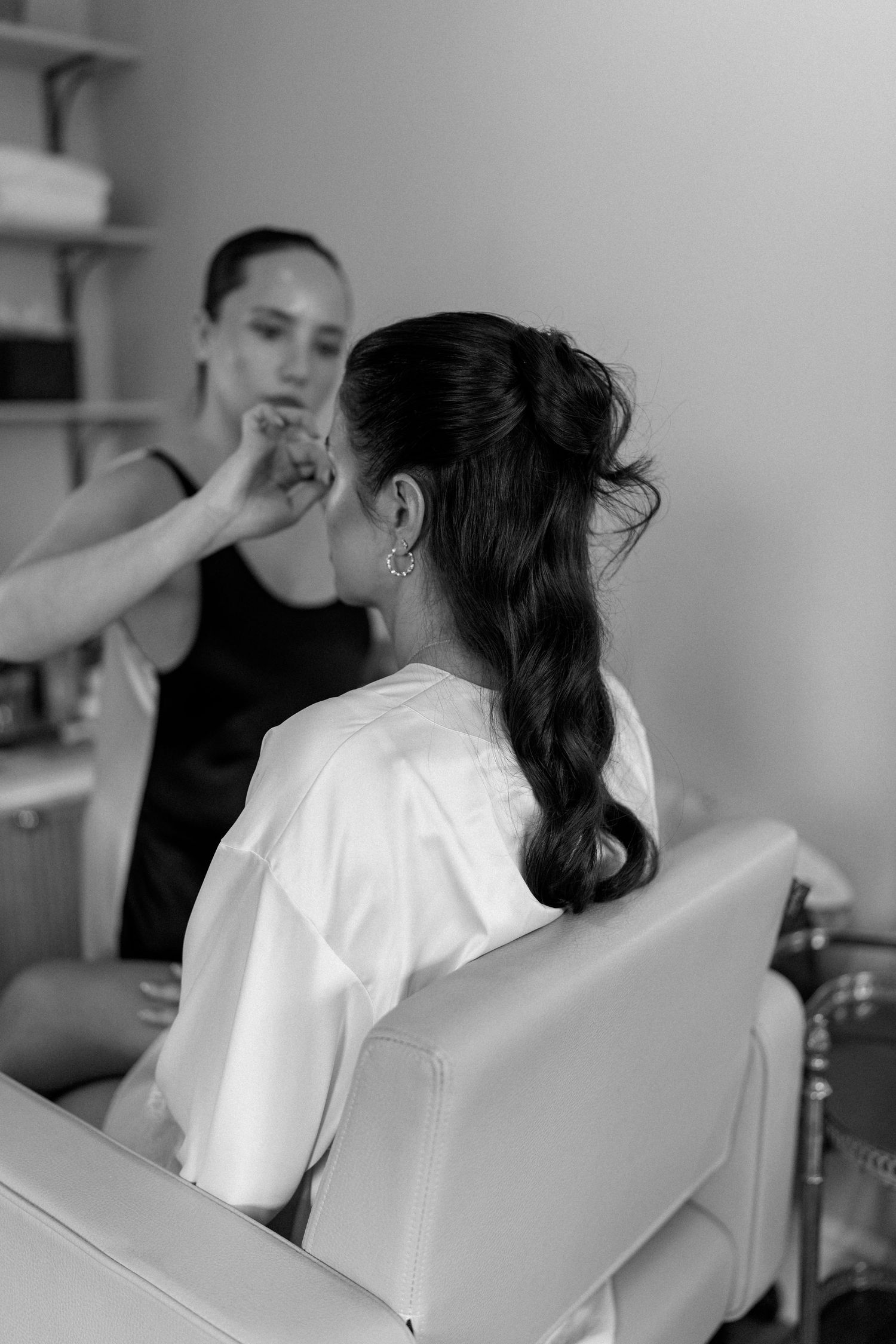 Makeup artist applying makeup to dark-haired bride in white robe at salon chair, black and white photo.