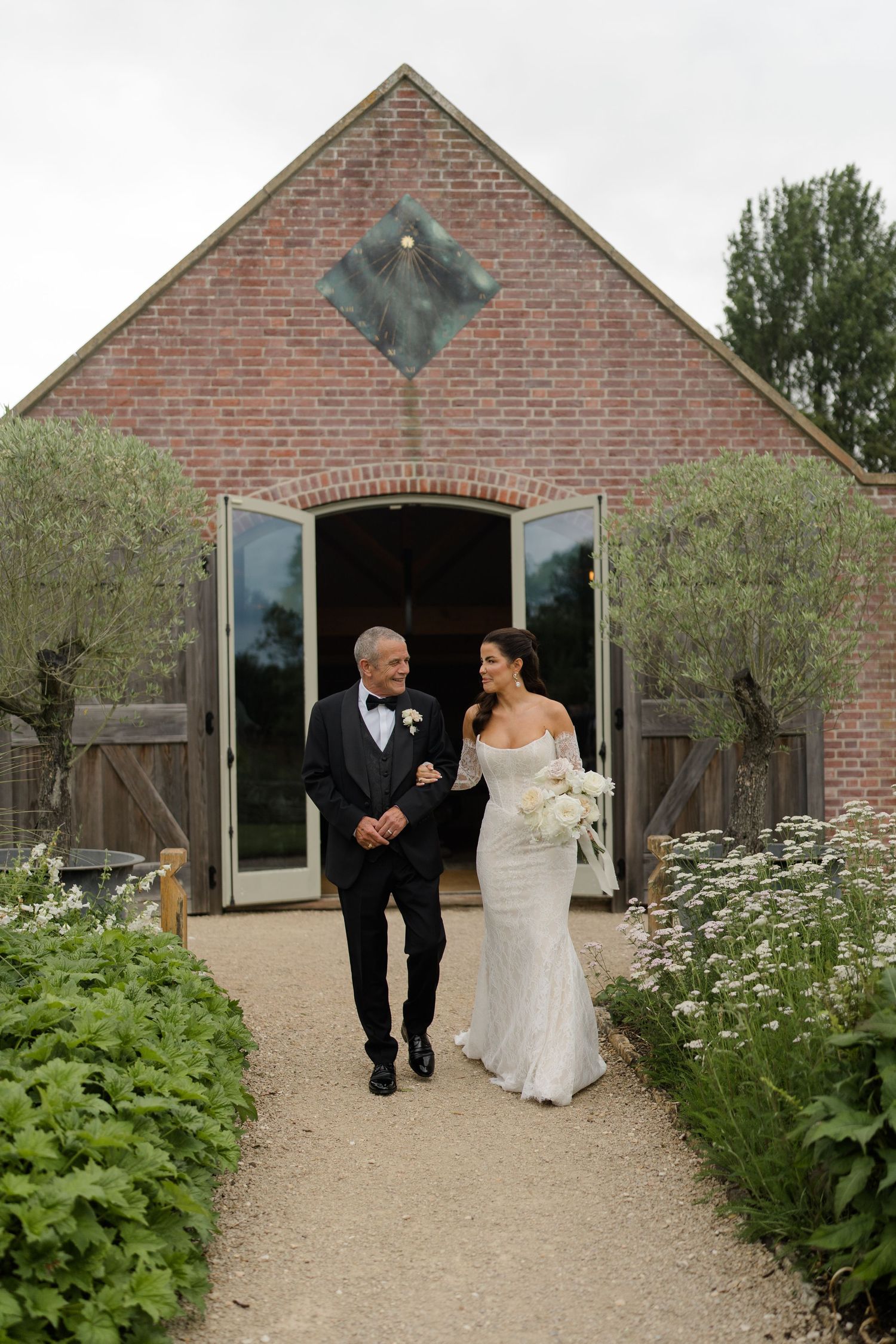Bride in fitted white lace gown walks with father from rustic brick barn along flower-lined gravel path.