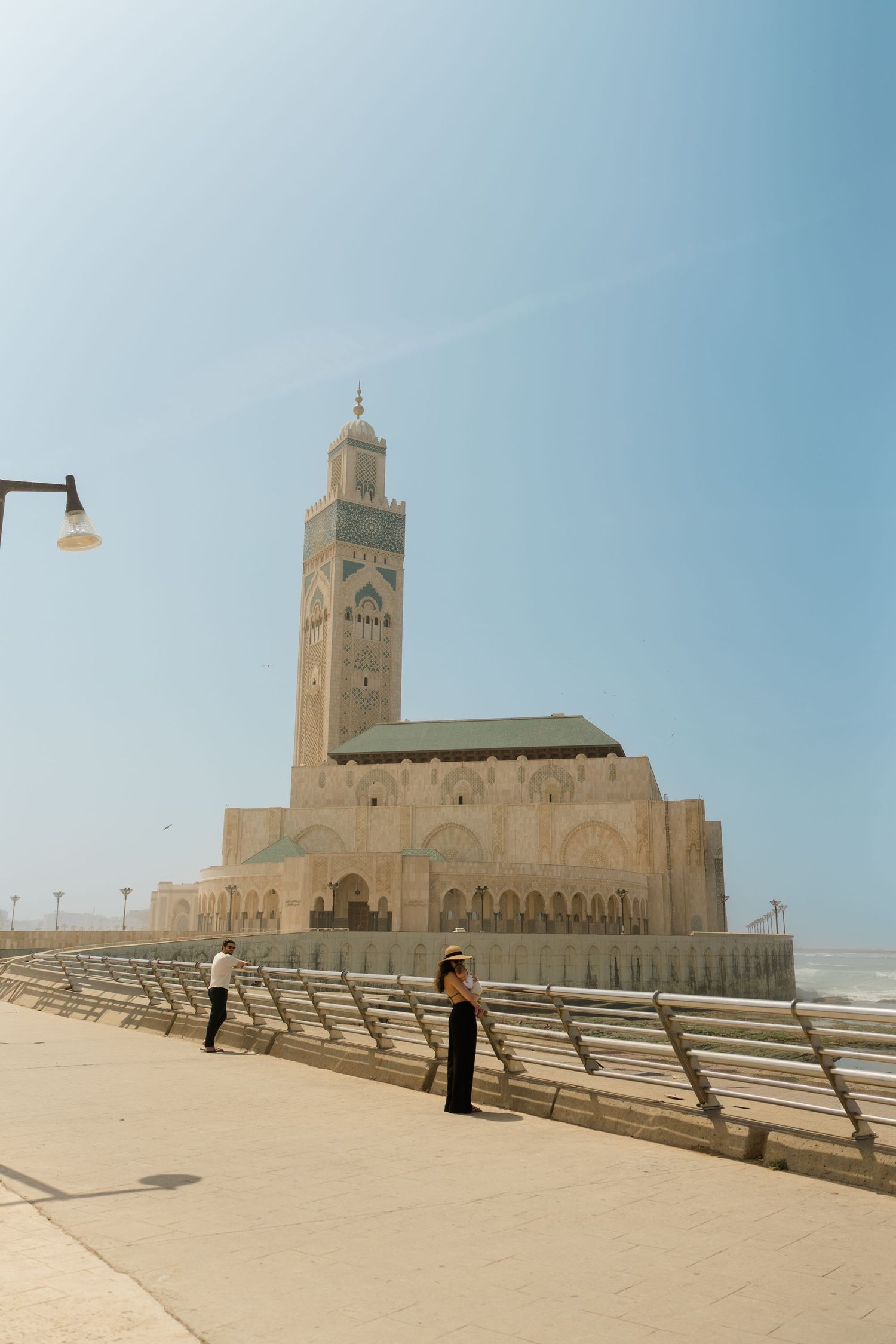 Tourists walk along the promenade near the iconic Hassan II Mosque in Casablanca, Morocco, under a clear blue sky.