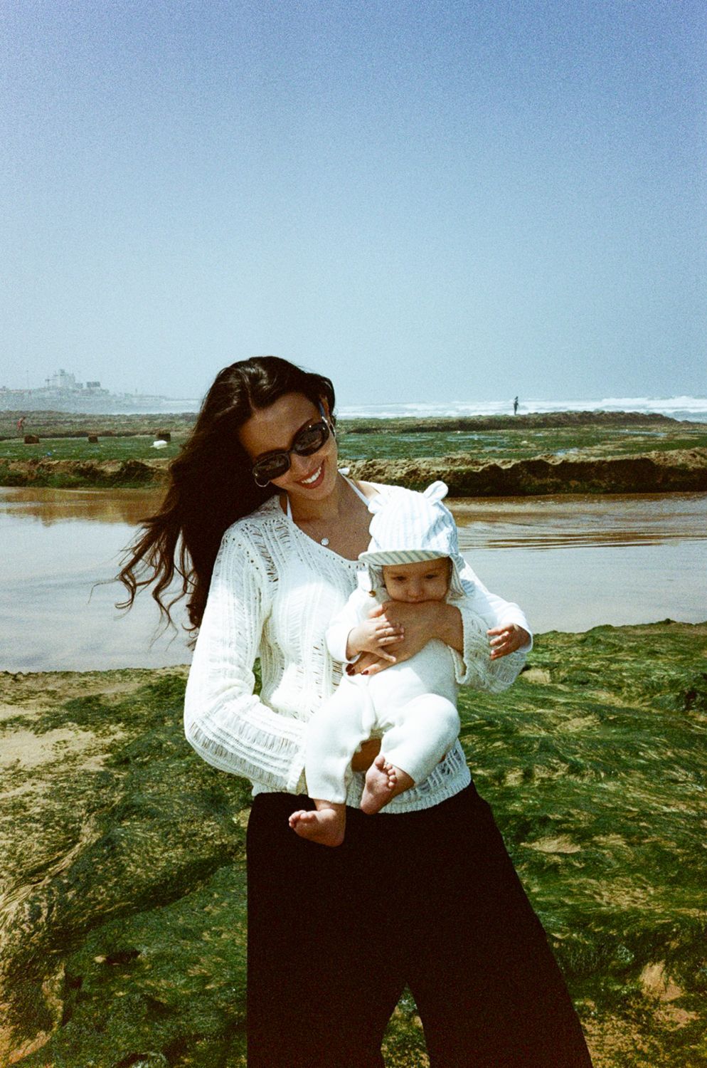A smiling woman in white lace top and sunglasses holds a baby near a rocky beach on a sunny day.