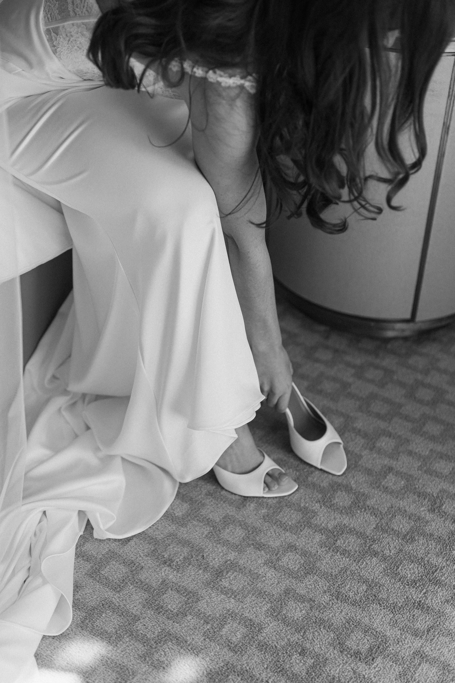 Black and white photo of a bride in a white gown putting on open-toe white bridal heels before her wedding.