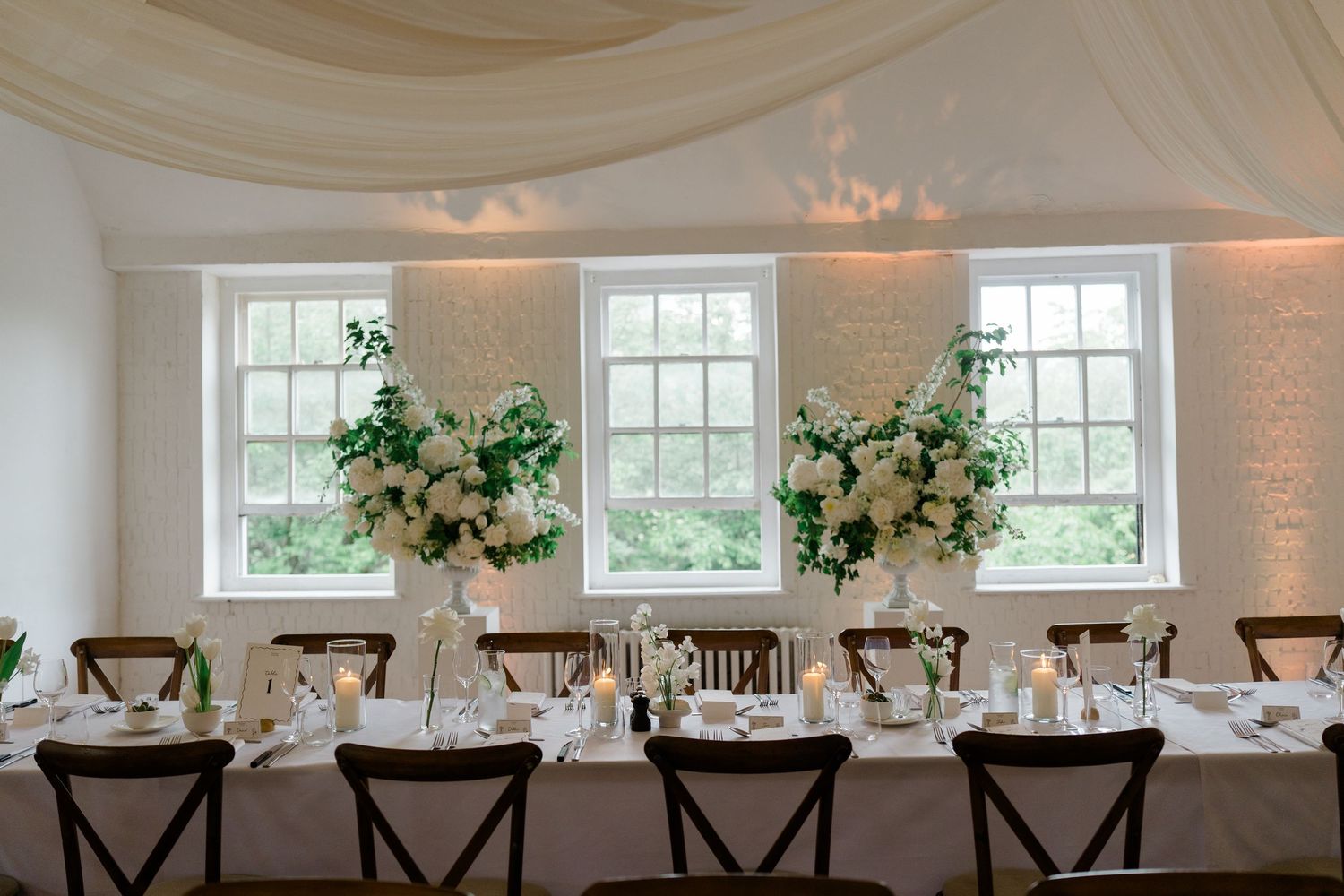 Elegant wedding reception head table with tall white floral centerpieces and candlelight by windows.