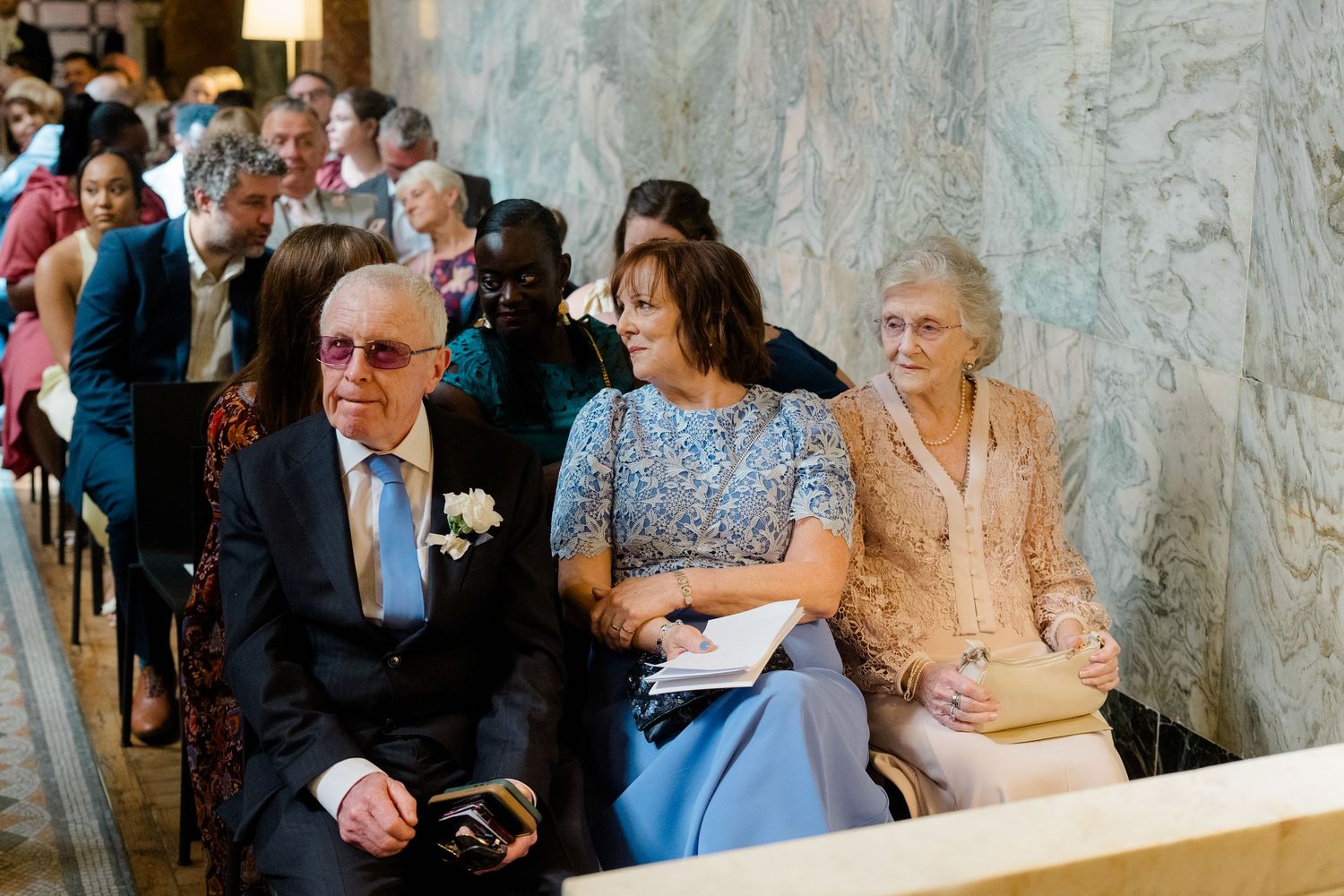 Elderly guests seated in rows at a wedding ceremony, dressed in formal attire inside an elegant marble venue.