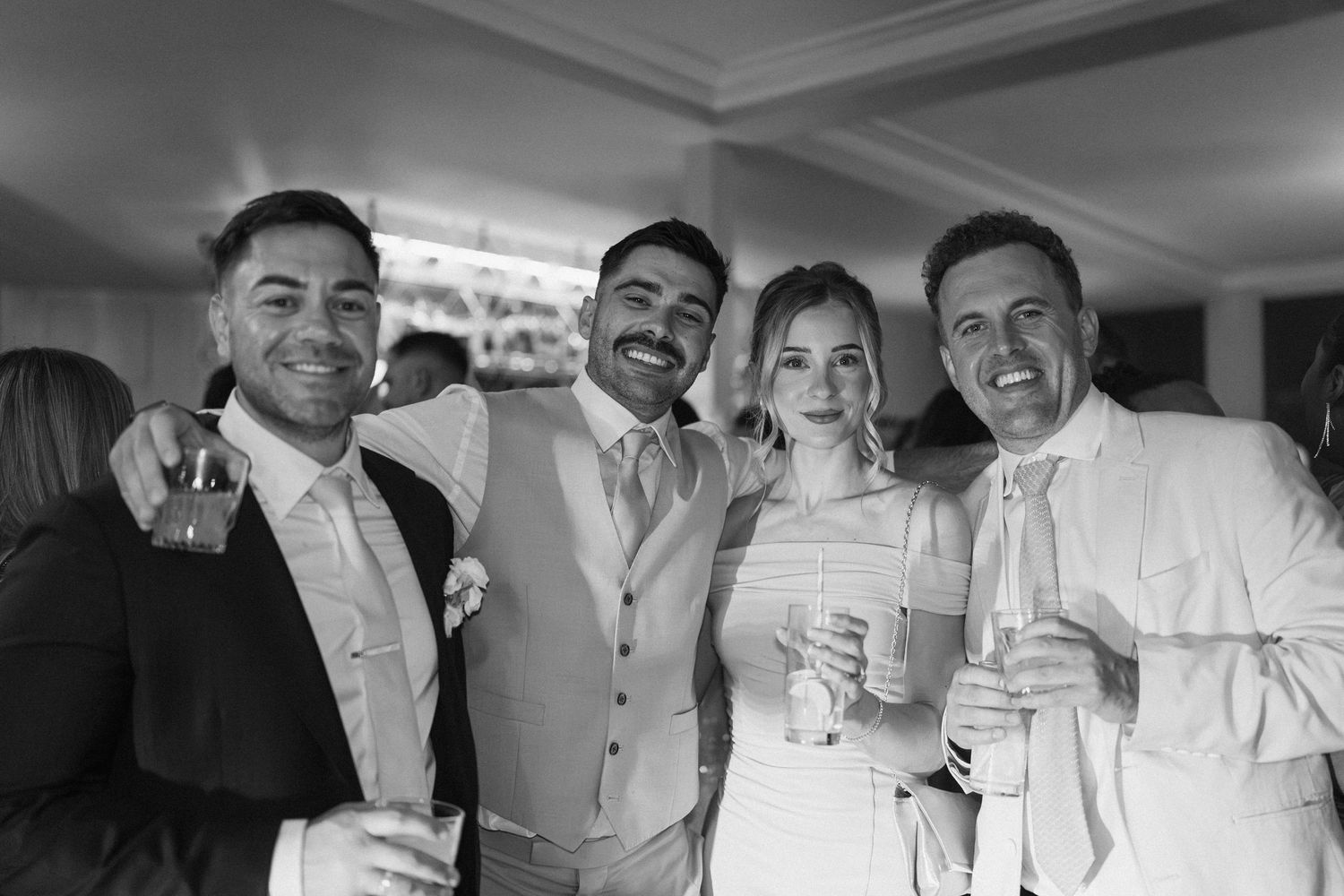 Black and white photo of four smiling wedding guests posing together holding drinks at reception.