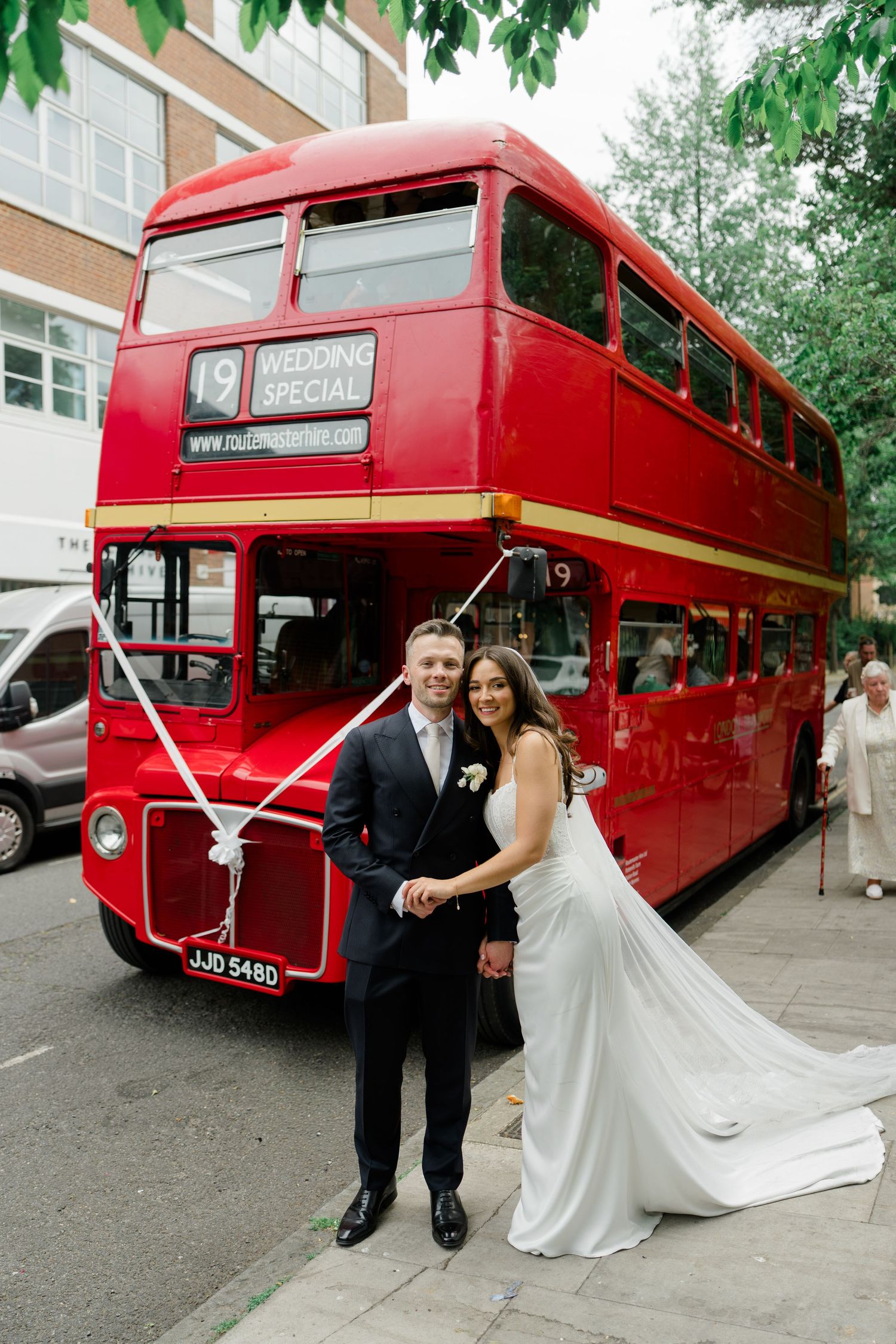 Smiling bride and groom posing in front of a classic red London double-decker wedding bus.
