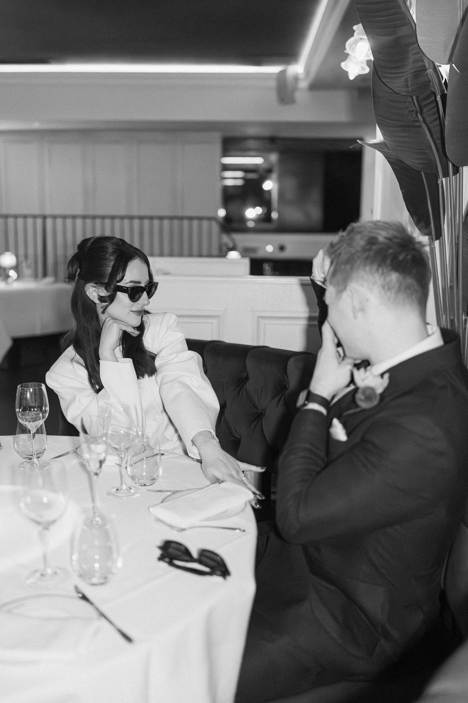 Elegant couple in black and white photo dining at restaurant table with wine glasses, woman wearing sunglasses.