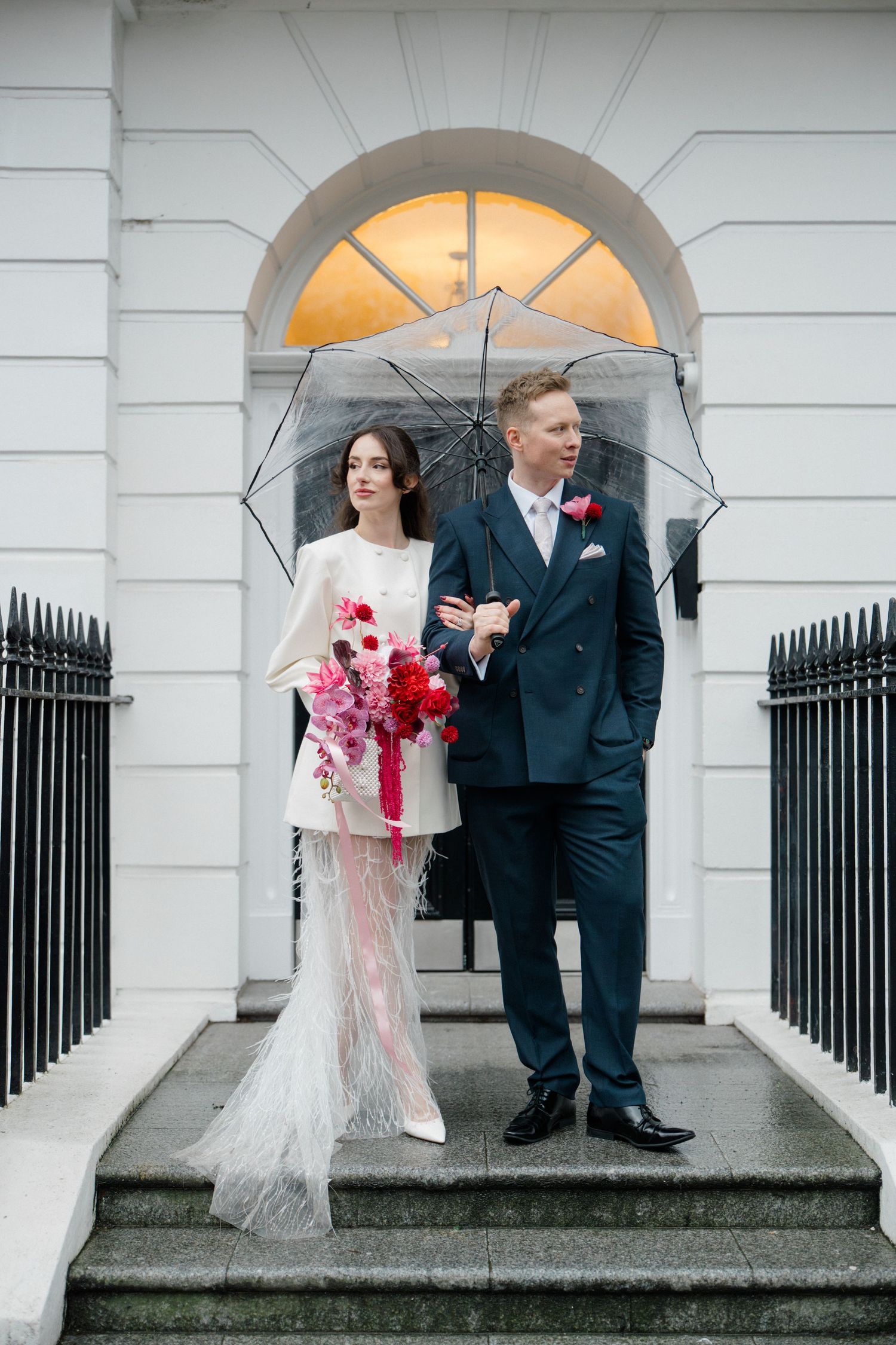 Bride and groom standing on steps under clear umbrella, bride holding vibrant pink and red bouquet.