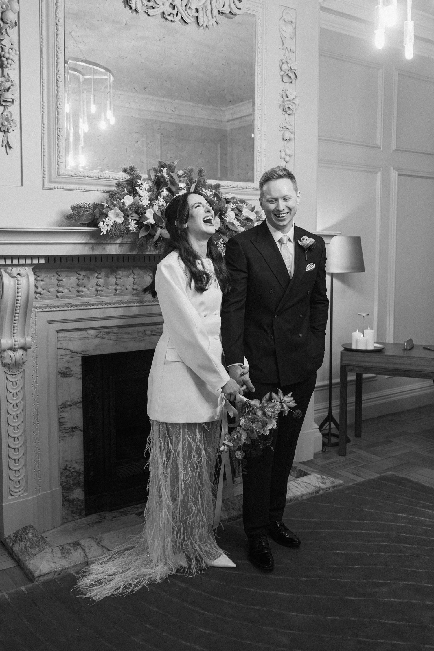 Bride and groom laughing joyfully before ornate fireplace with floral arrangement in black and white.