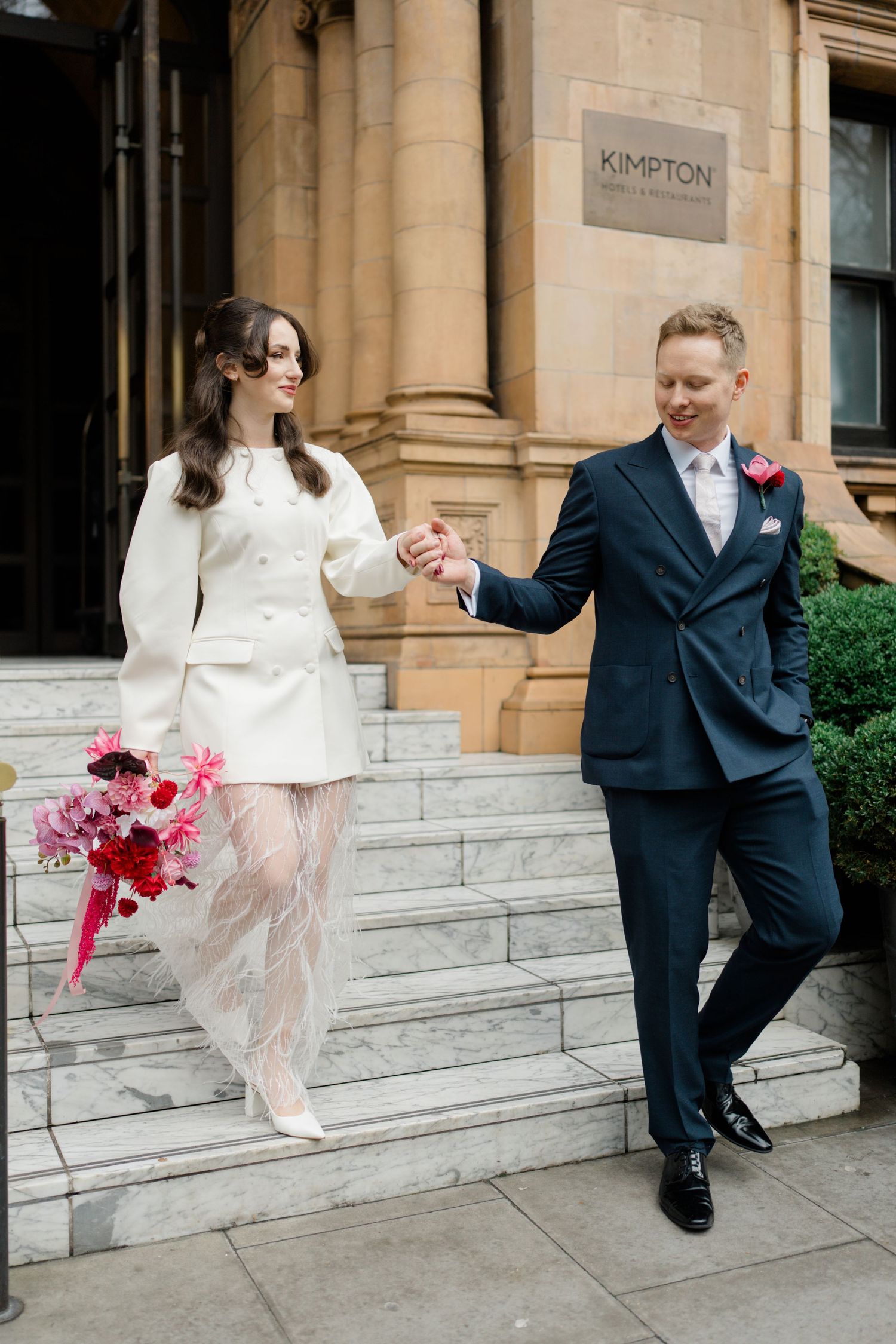 Bride in white jacket dress and groom in navy suit holding hands on stone steps outside Kimpton Hotel.