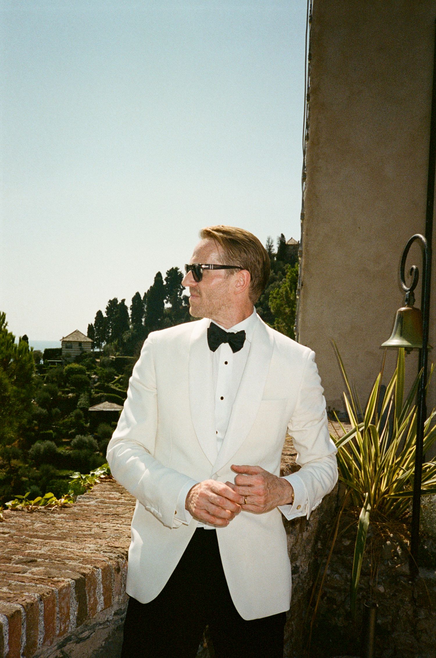 A man in a white tuxedo jacket and black bow tie poses on a sunny terrace with lush greenery in the background.