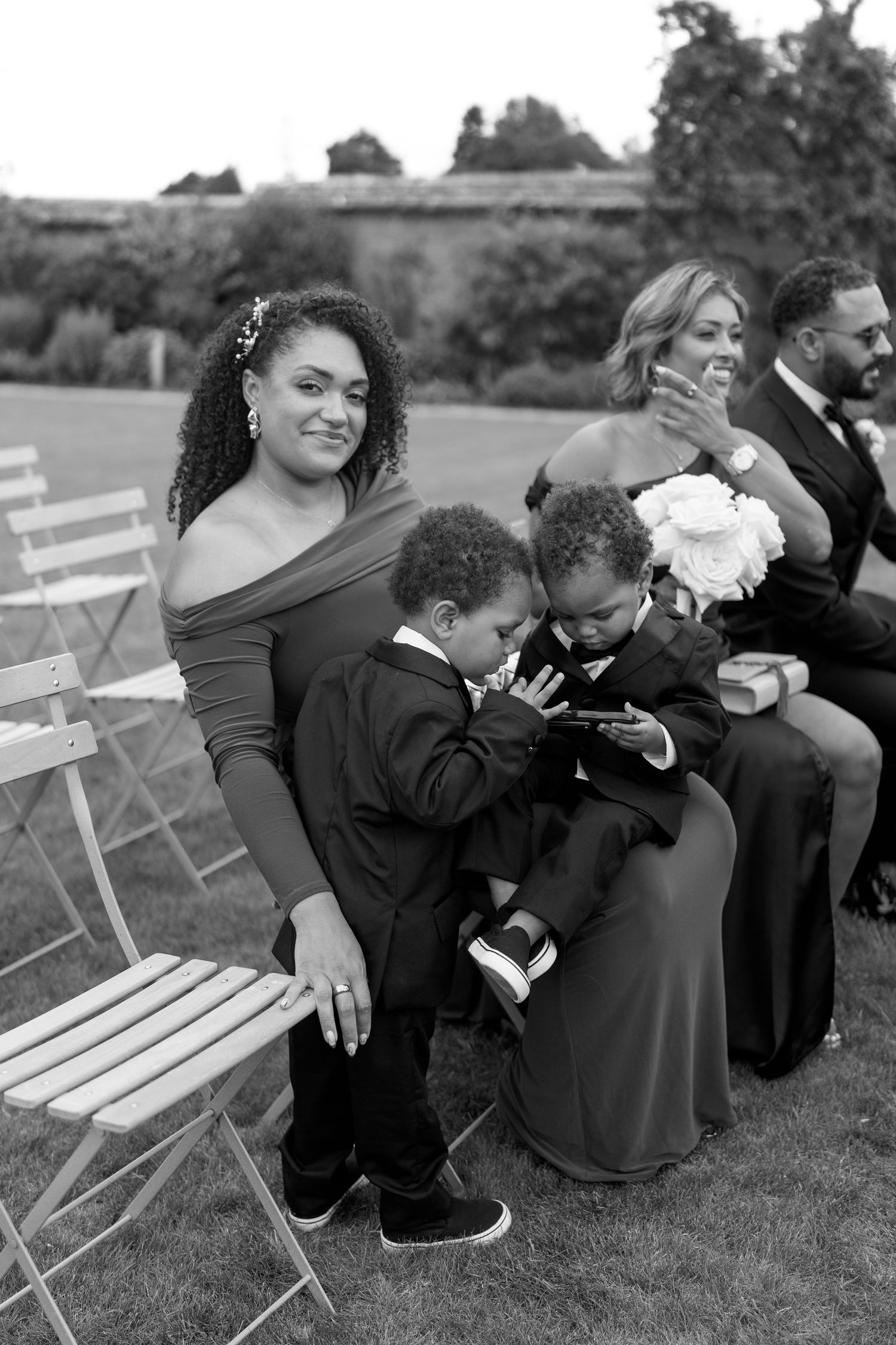 Woman with two young boys in tuxedos seated at an outdoor wedding ceremony in black and white.