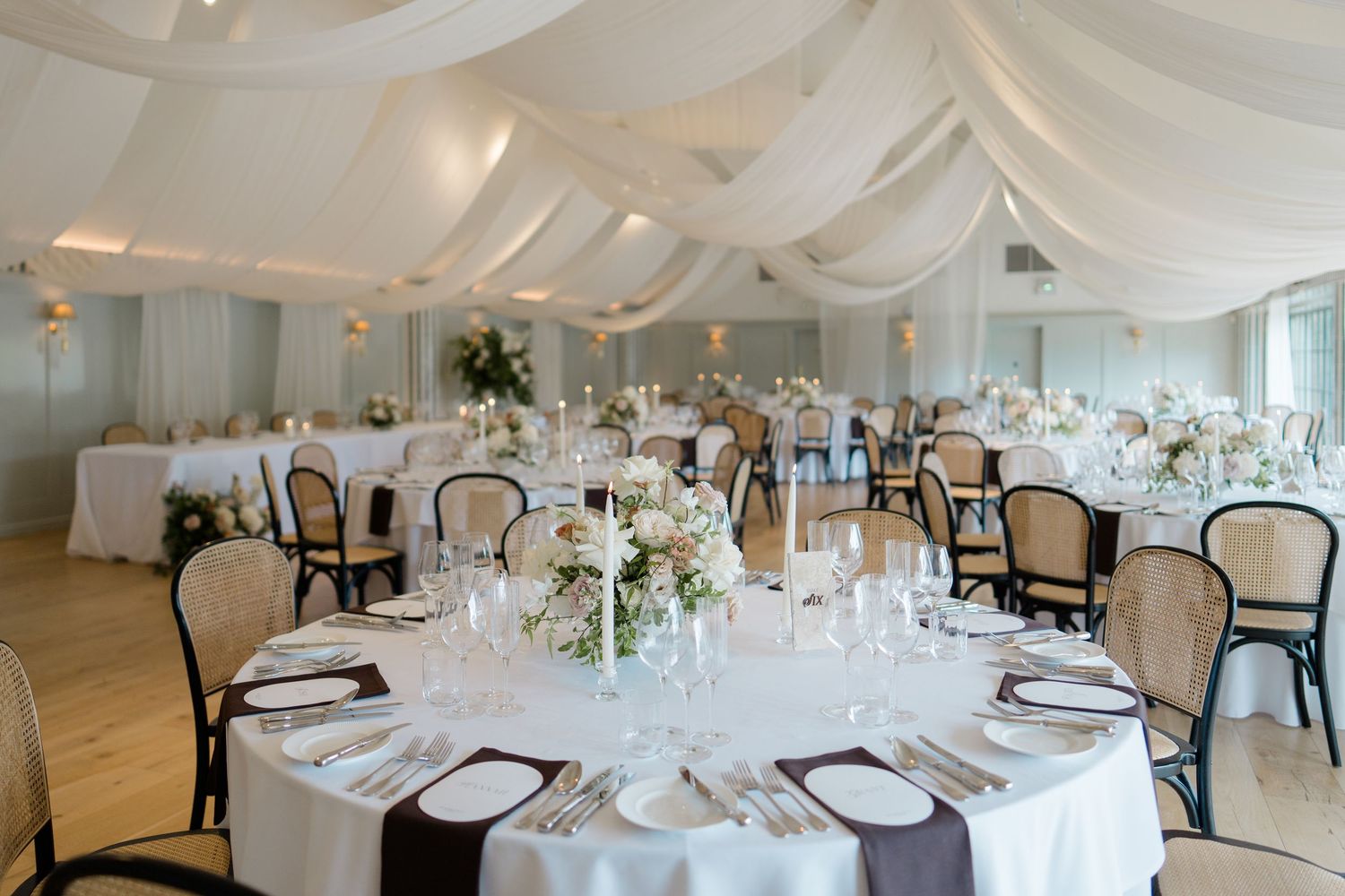 Elegant wedding reception hall with white draped ceiling, round tables, and floral centerpieces.