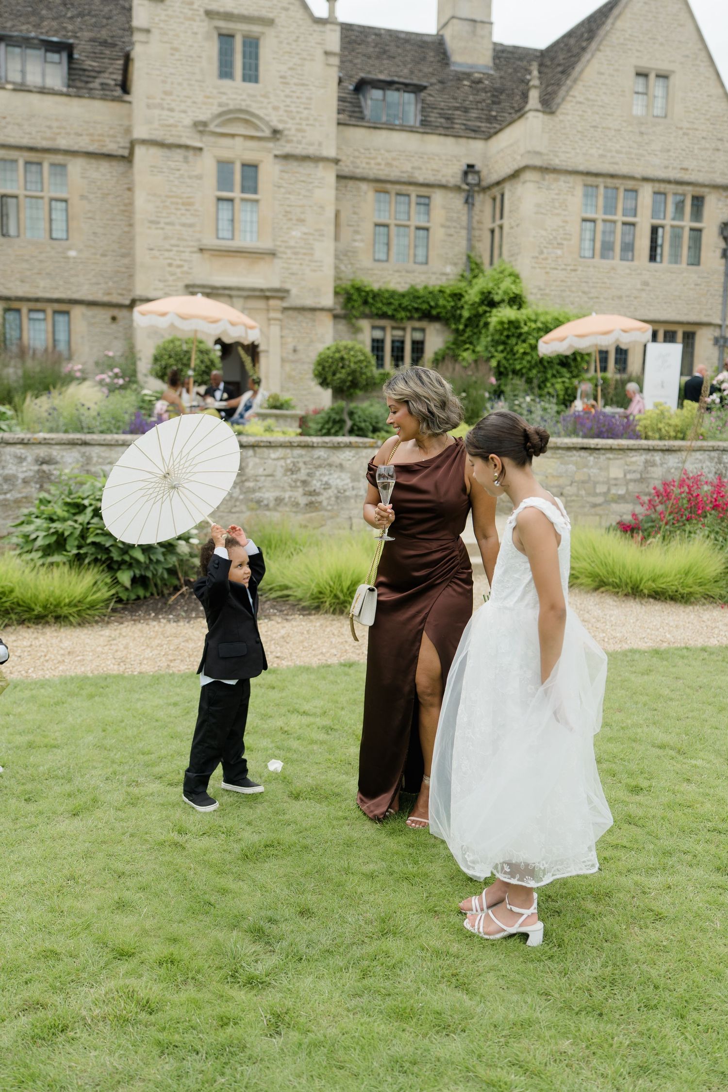 Woman in brown gown with children holding parasol on lawn outside grand stone manor wedding venue.