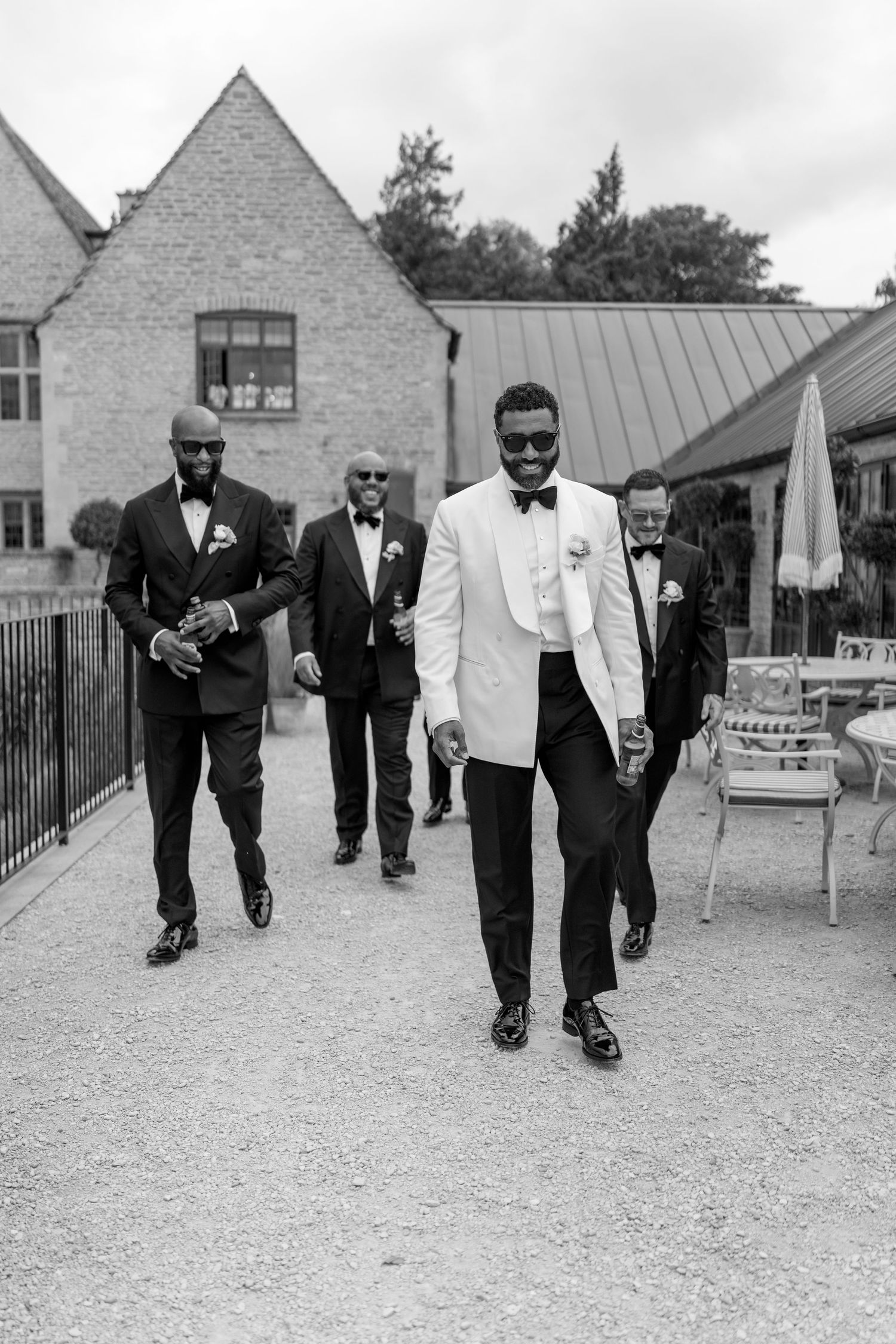 Black and white photo of groom in white tuxedo jacket leading groomsmen across venue courtyard.
