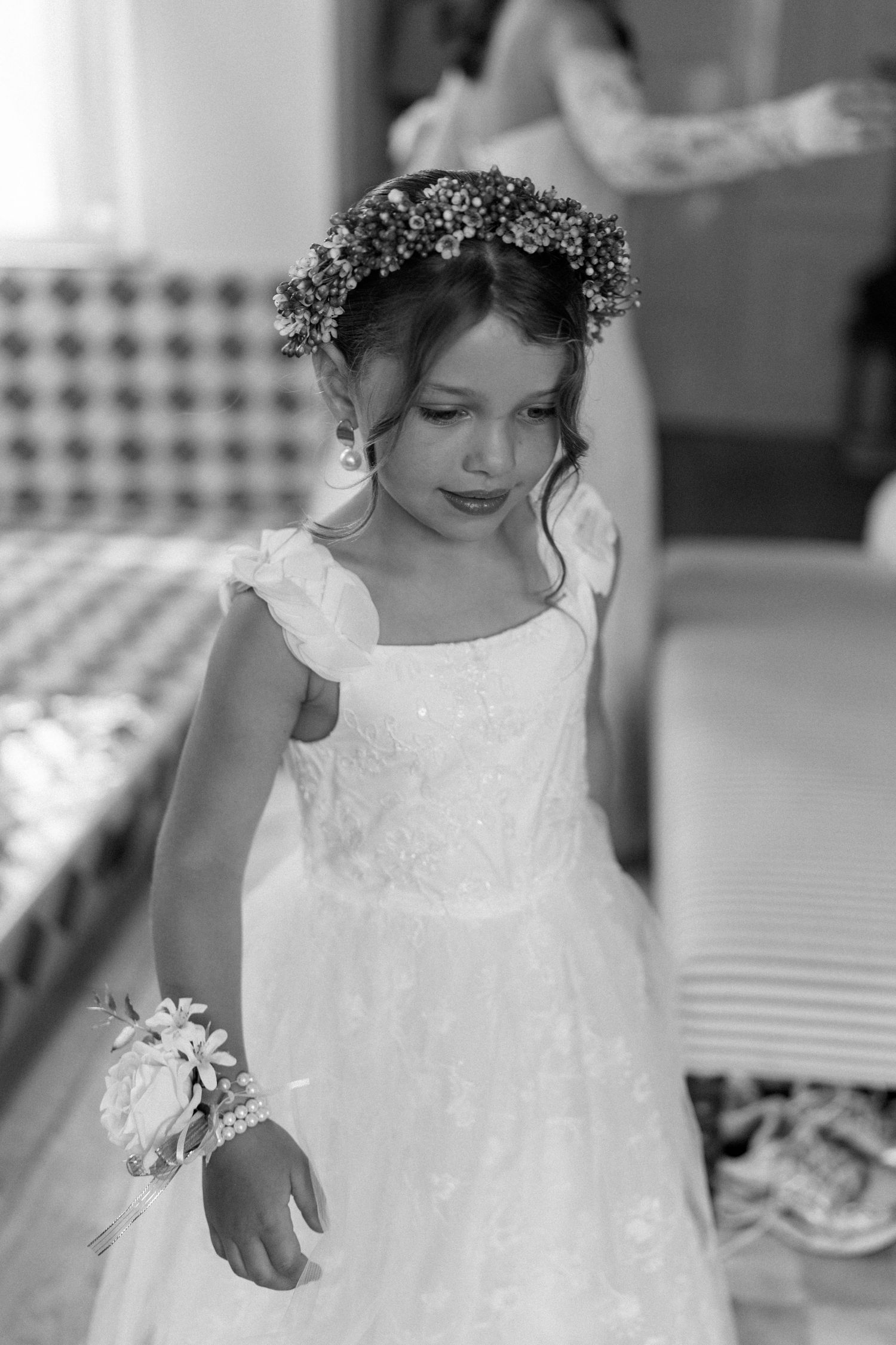 Young flower girl in white lace dress wearing floral crown and wrist corsage at wedding ceremony.