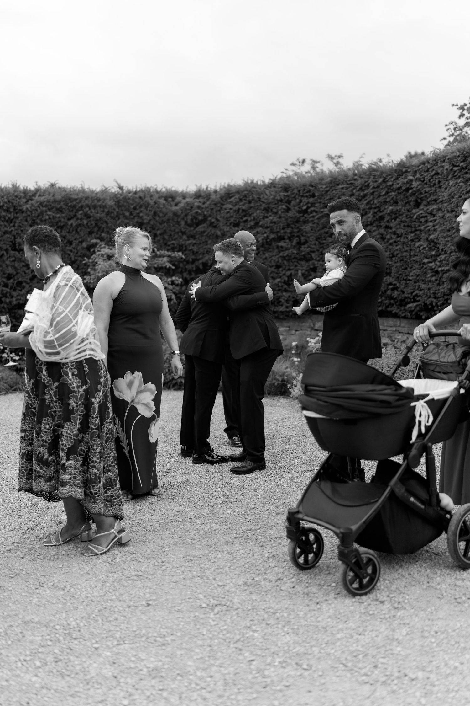 Wedding guests socializing outdoors in black and white photo, with baby and stroller near hedge backdrop.