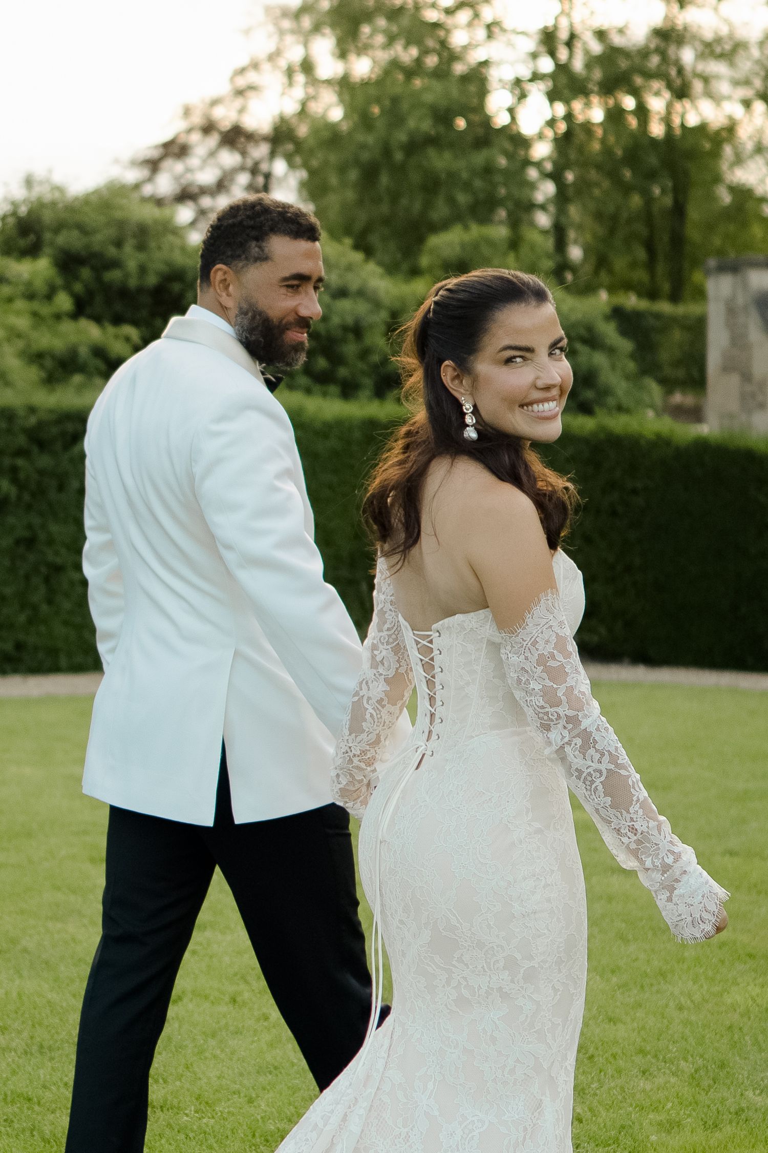 Smiling bride in lace off-shoulder gown glances back while walking with groom in white tuxedo jacket.