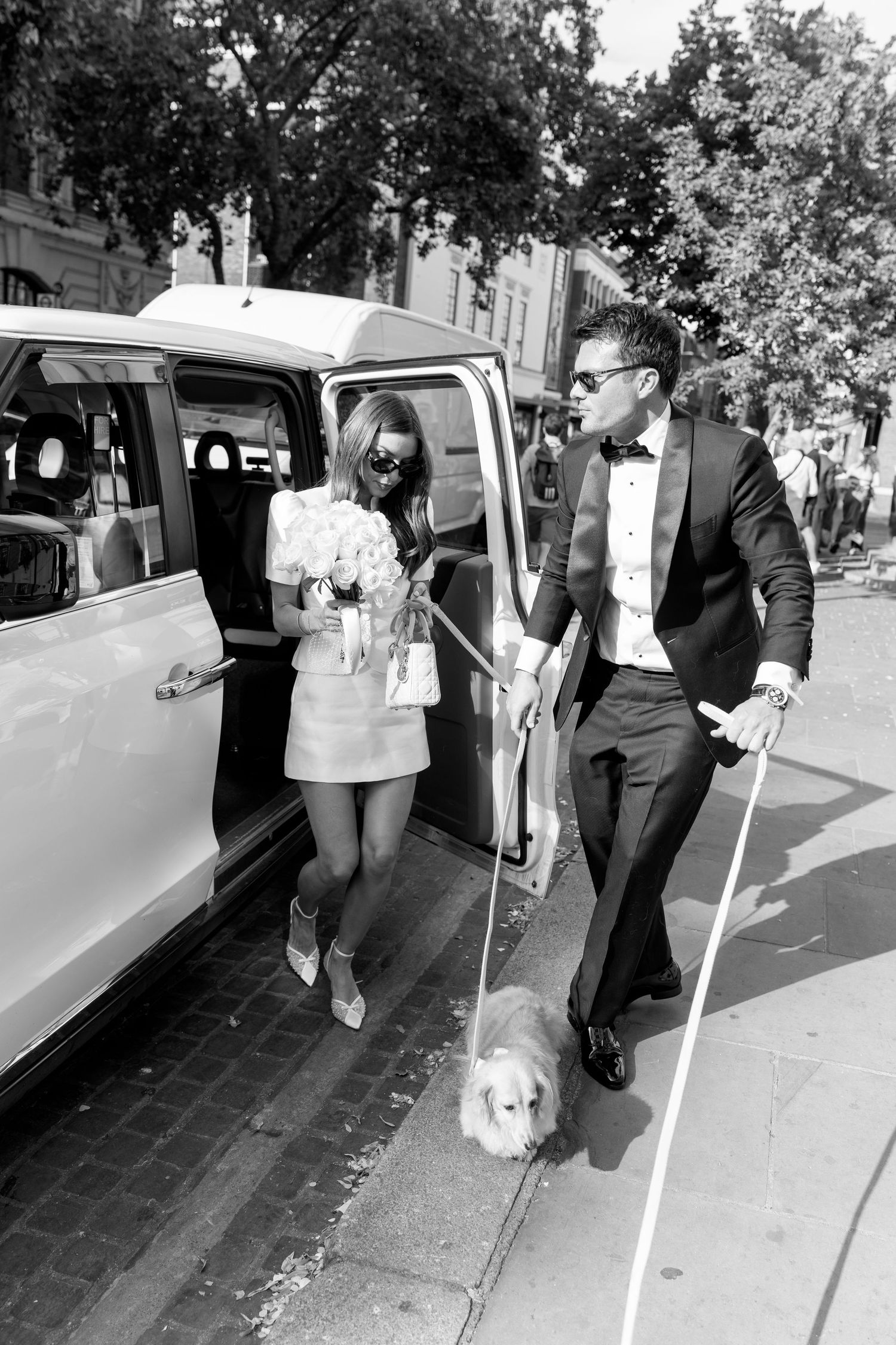 Black and white photo of bride and groom with corgi exiting a white vintage car on a city street.