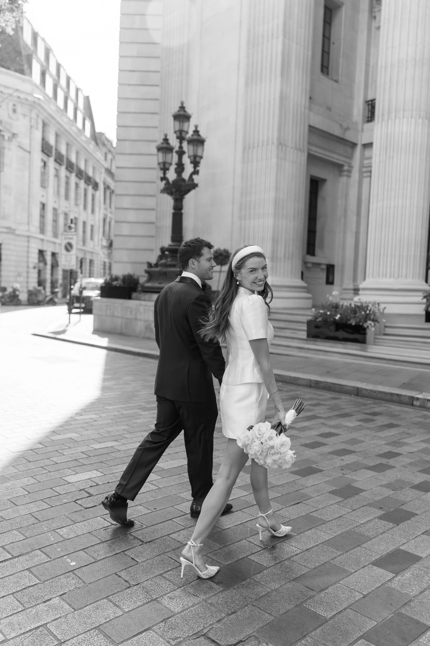Bride and groom walking hand-in-hand past grand neoclassical columns on sunny city street.