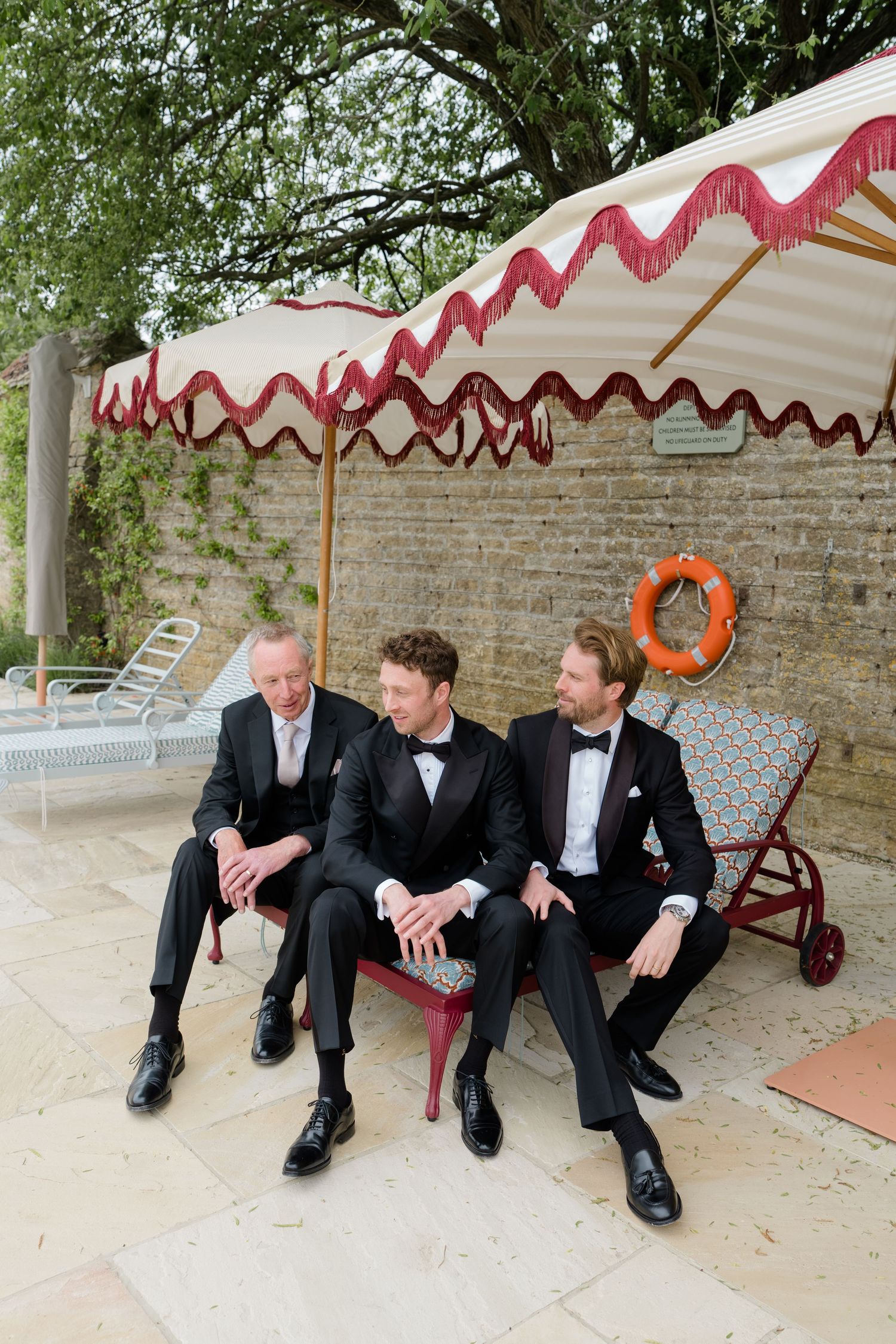 Three groomsmen in black tuxedos sitting under a striped fringed umbrella by a pool at a wedding.