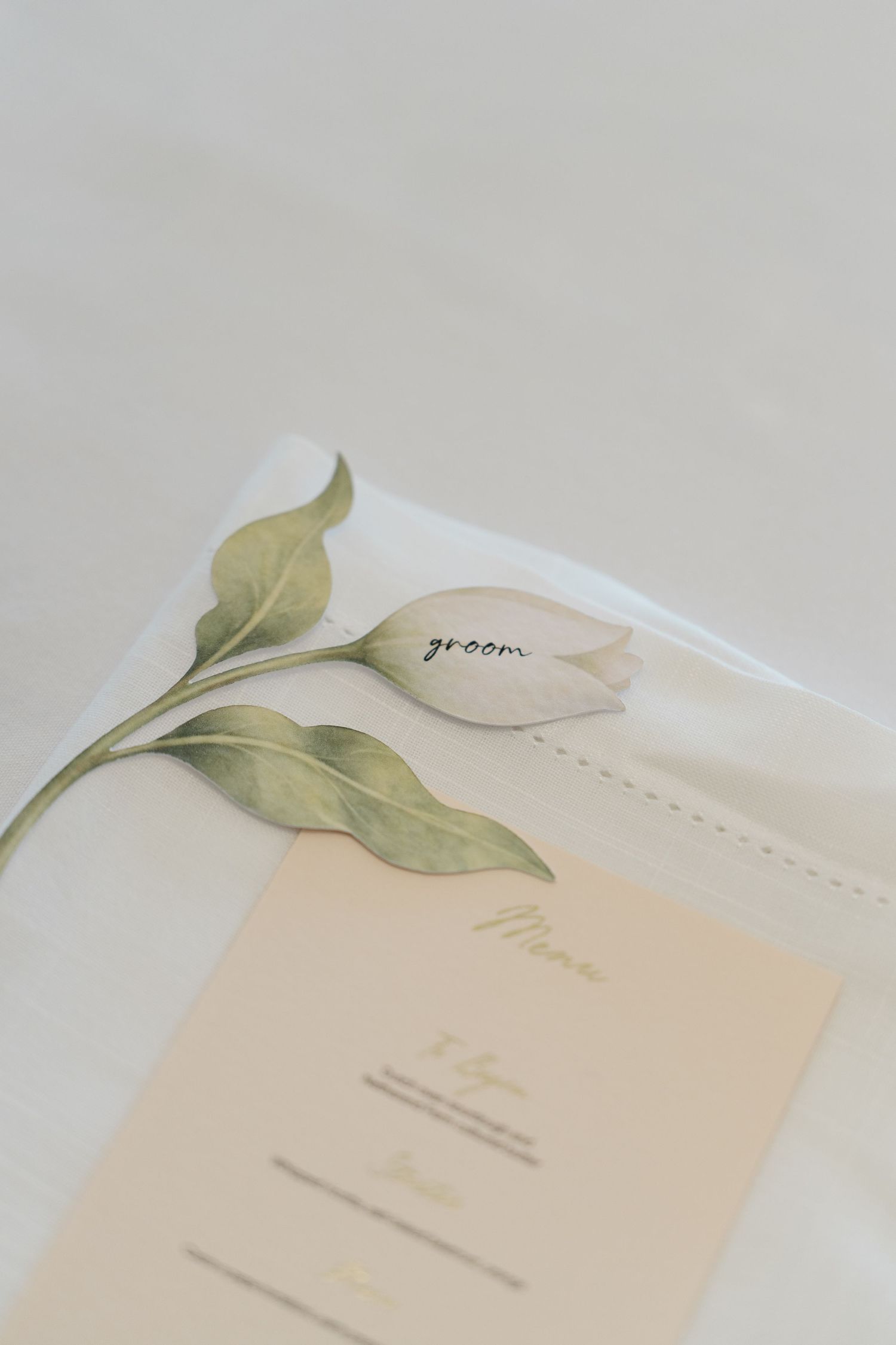 Wedding place card reading groom rests on illustrated tulip design atop white linen with cream menu card.
