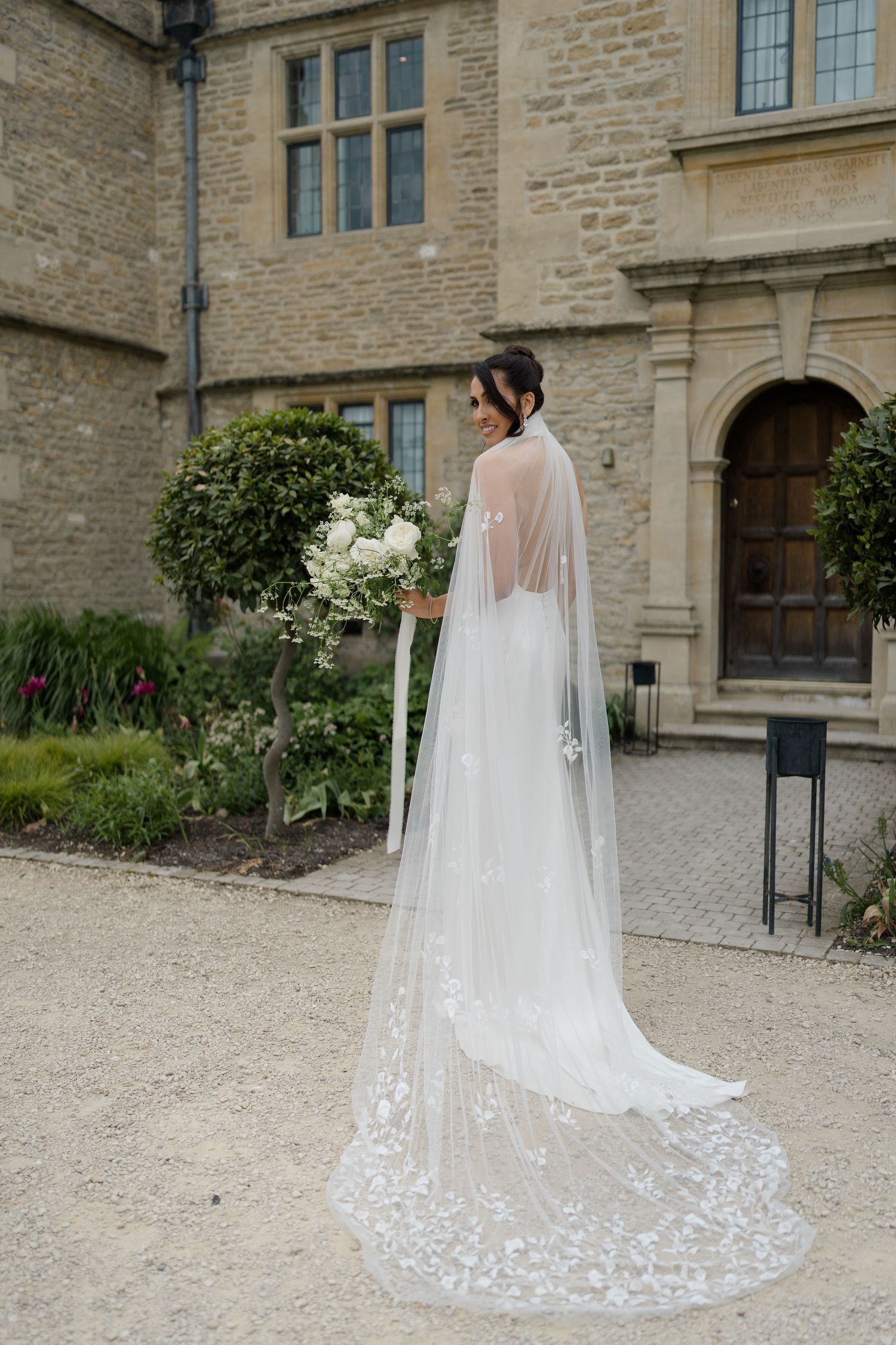 Bride in elegant white gown with lace cathedral veil stands outside historic stone manor holding white floral bouquet.