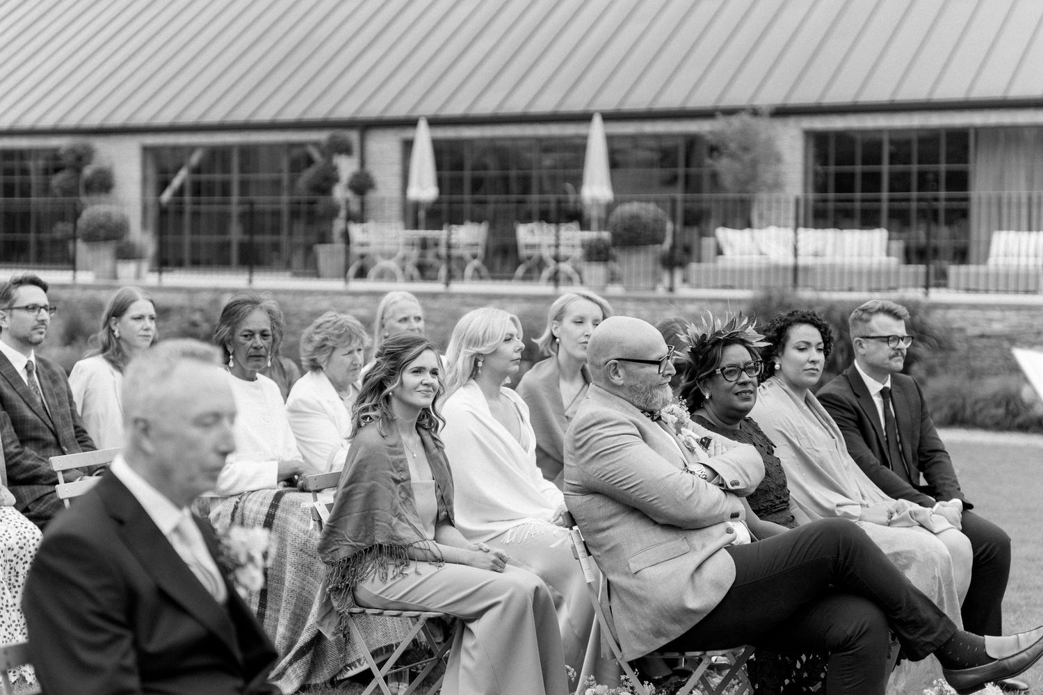 Black and white photo of elegantly dressed wedding guests seated in rows at an outdoor ceremony.
