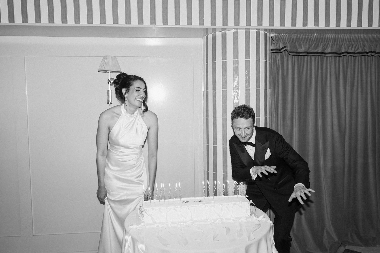 Black and white photo of laughing bride and groom leaning over champagne tower at wedding reception.