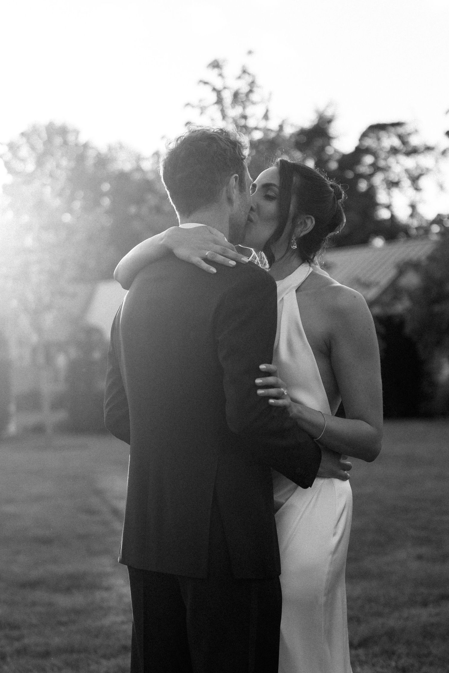 Black and white photo of a bride and groom sharing a kiss outdoors, with soft sunlight glowing behind them.