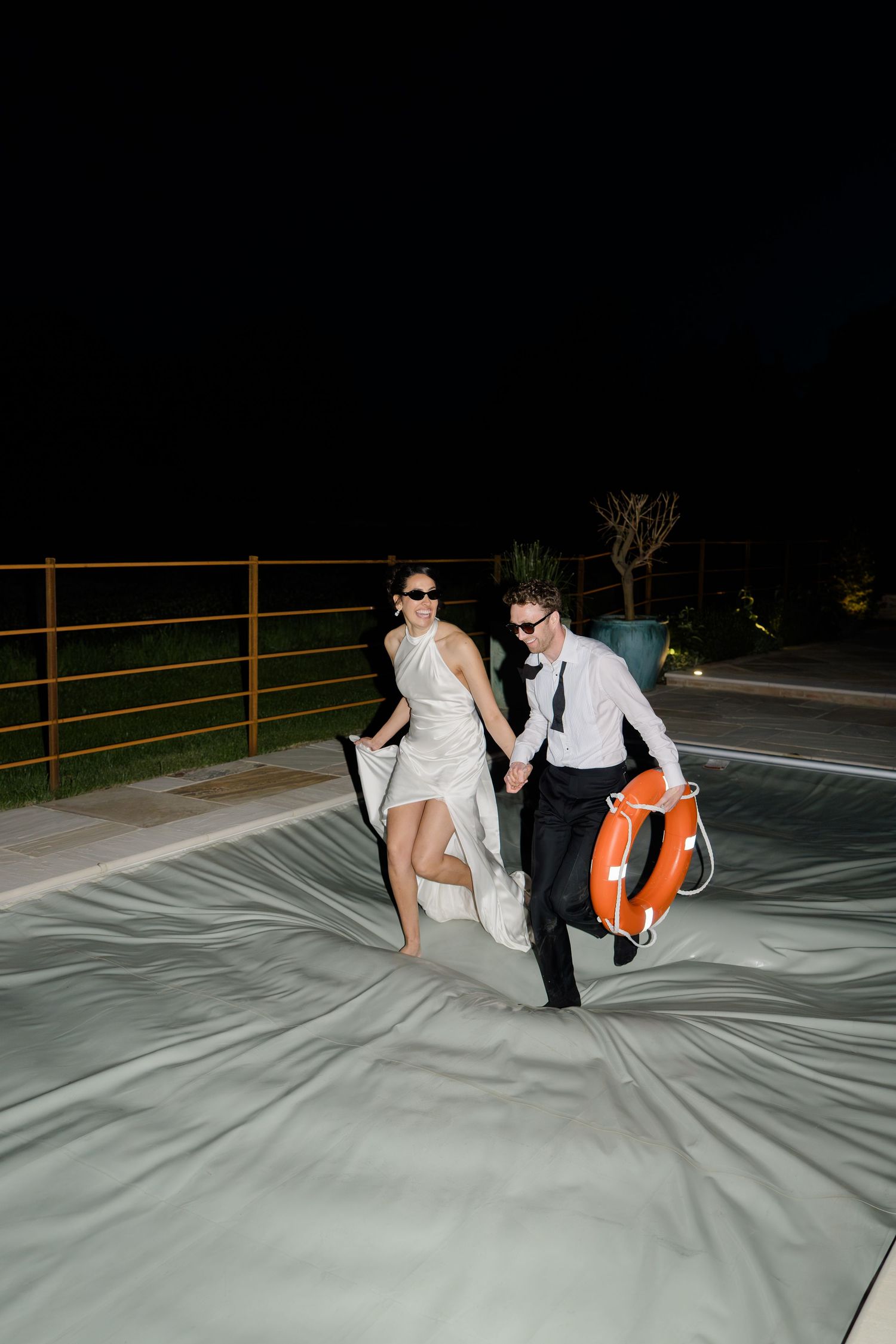 Laughing bride and groom run across covered pool at night holding a life preserver ring.