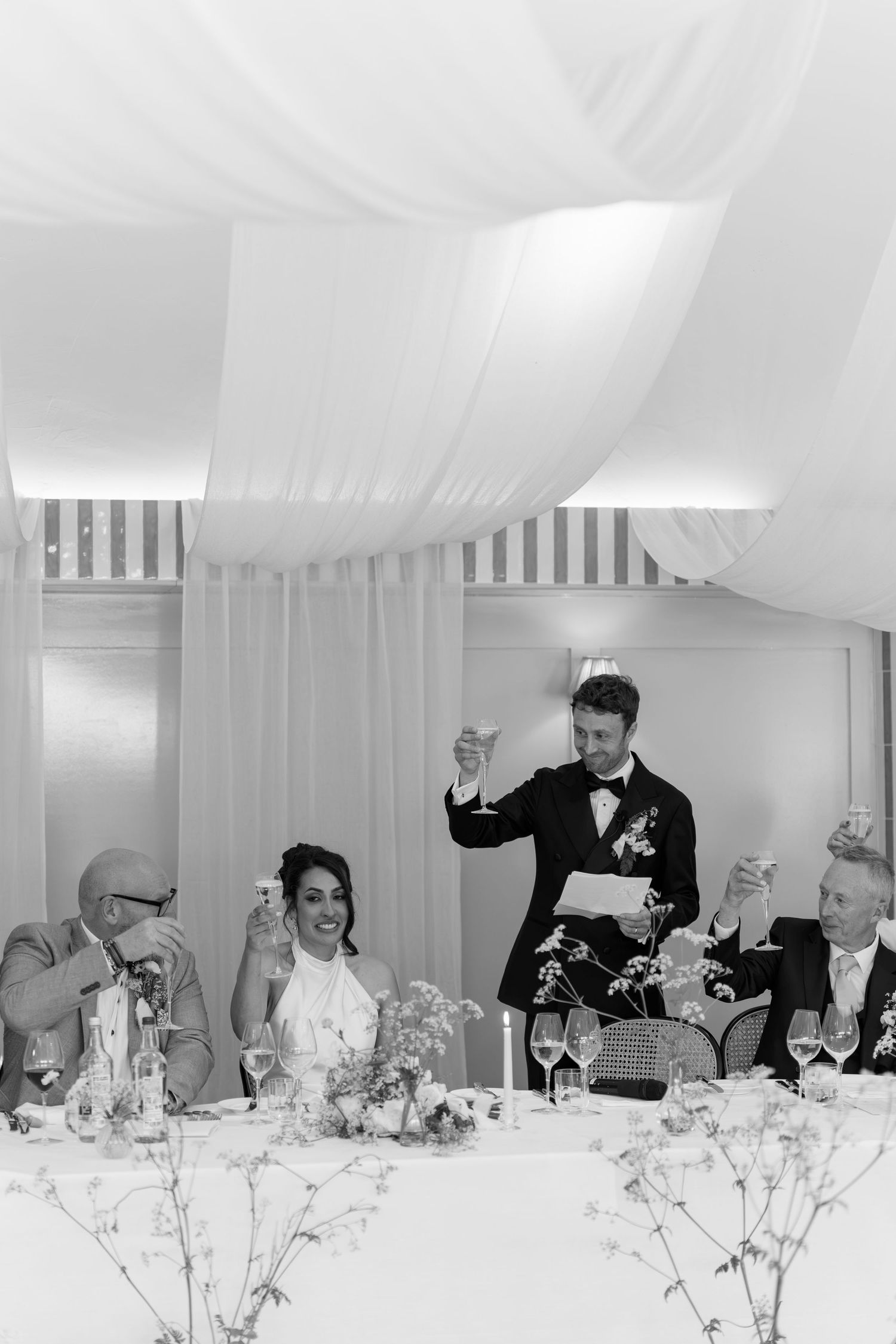 Black and white photo of a groom giving a wedding speech while raising a glass at the top table.