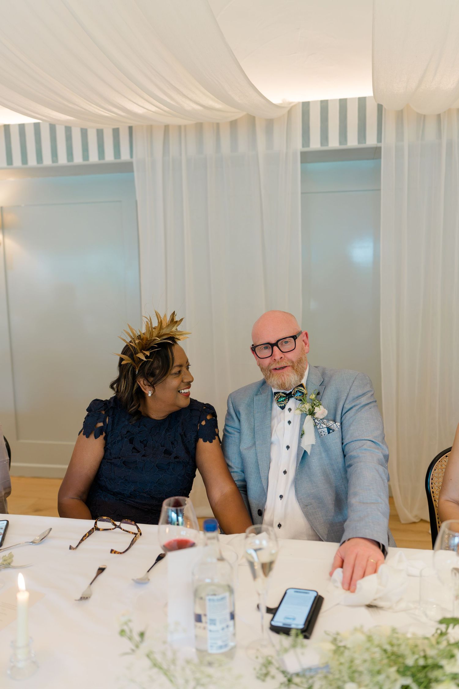 Smiling wedding guests in navy and light blue outfits seated at reception dinner table.