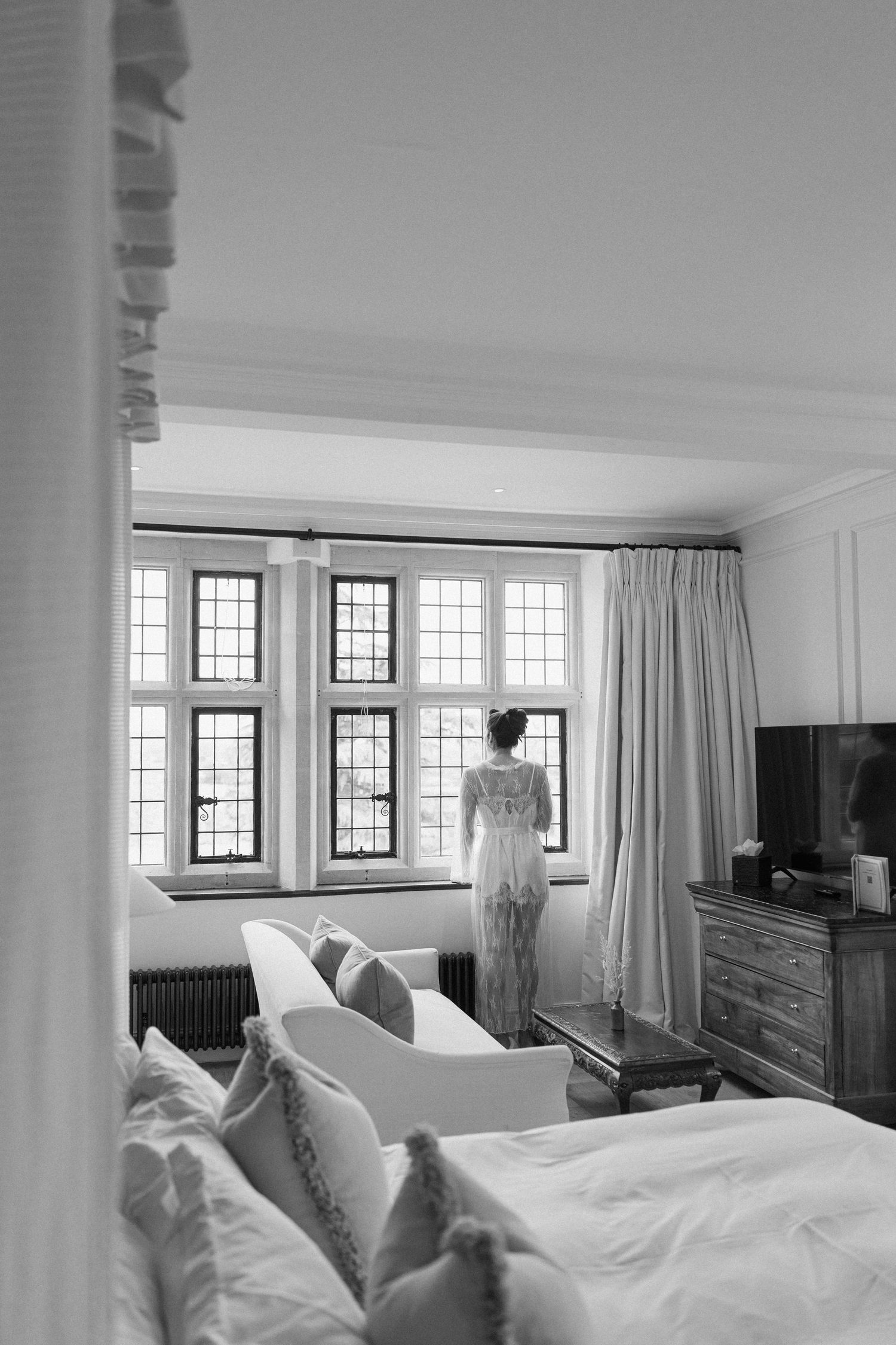 Black and white photo of bride in lace robe standing before large windows in an elegant hotel bedroom.