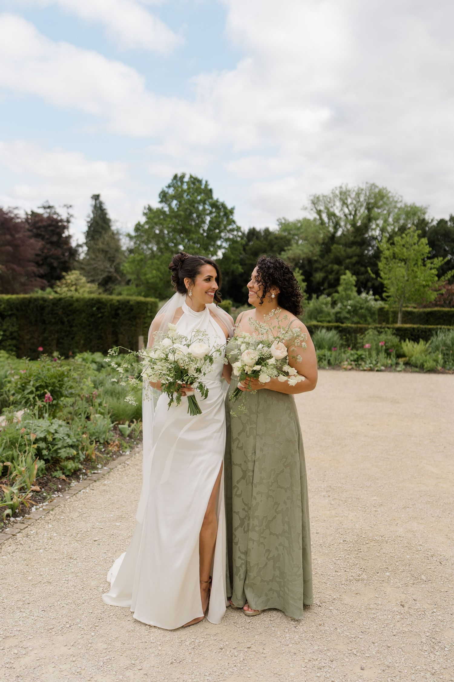 Bride in white gown and bridesmaid in sage green dress holding white bouquets in garden.