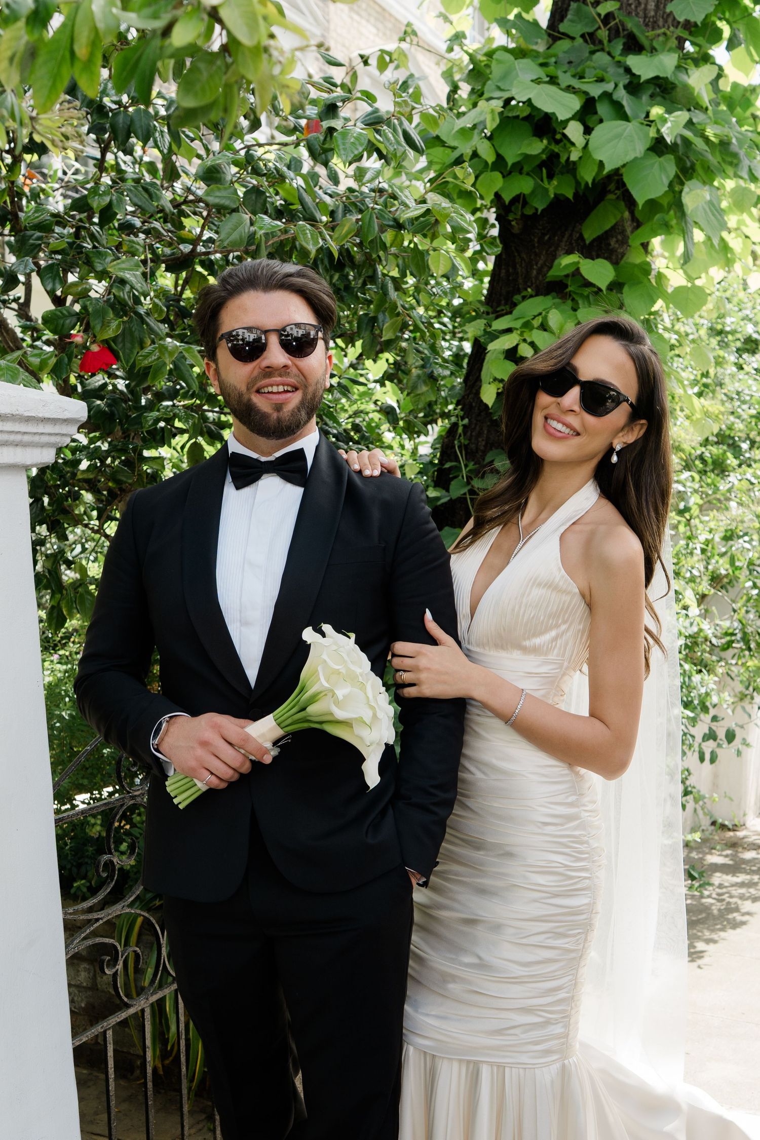 Stylish bride and groom in sunglasses posing outdoors, groom in black tuxedo holding white calla lily bouquet.