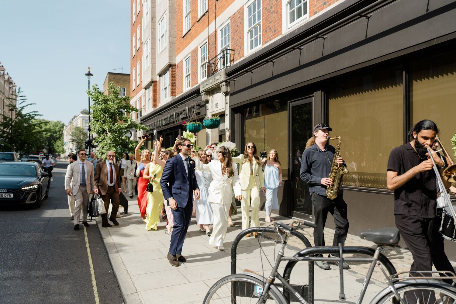 Wedding procession walking down a sunny London street with a saxophonist leading the celebration.