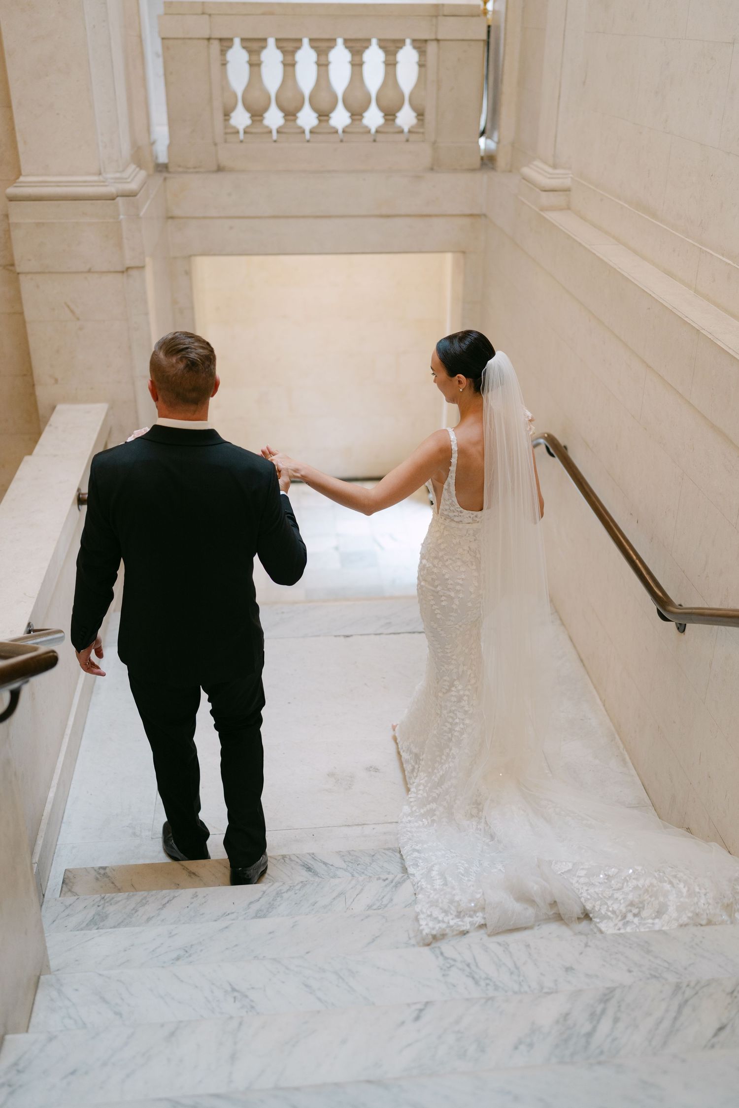 Couple descends marble staircase with bride's flowing lace train trailing behind.