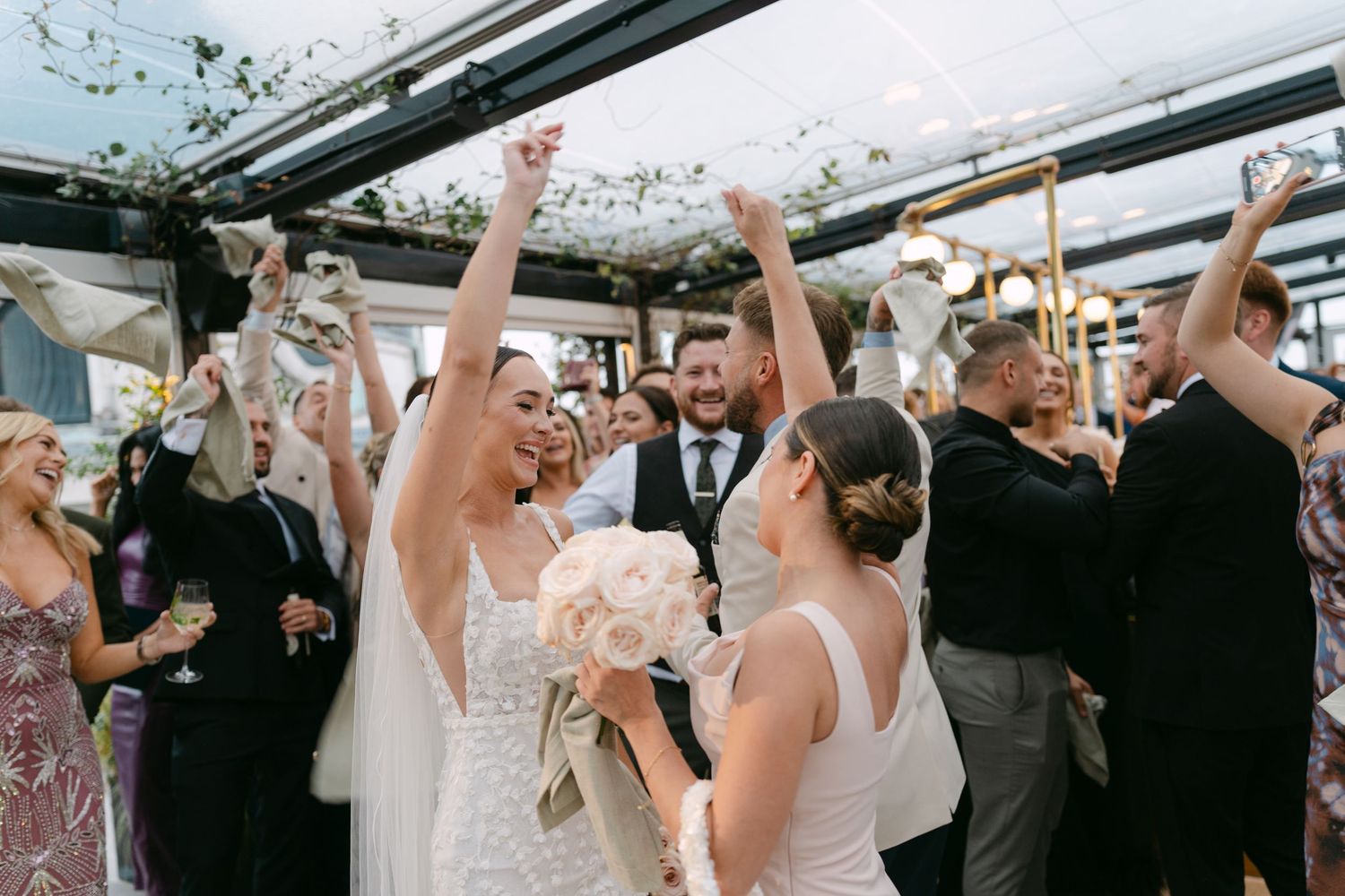 Wedding reception crowd raising their hands in celebration under a glass ceiling with hanging greenery.