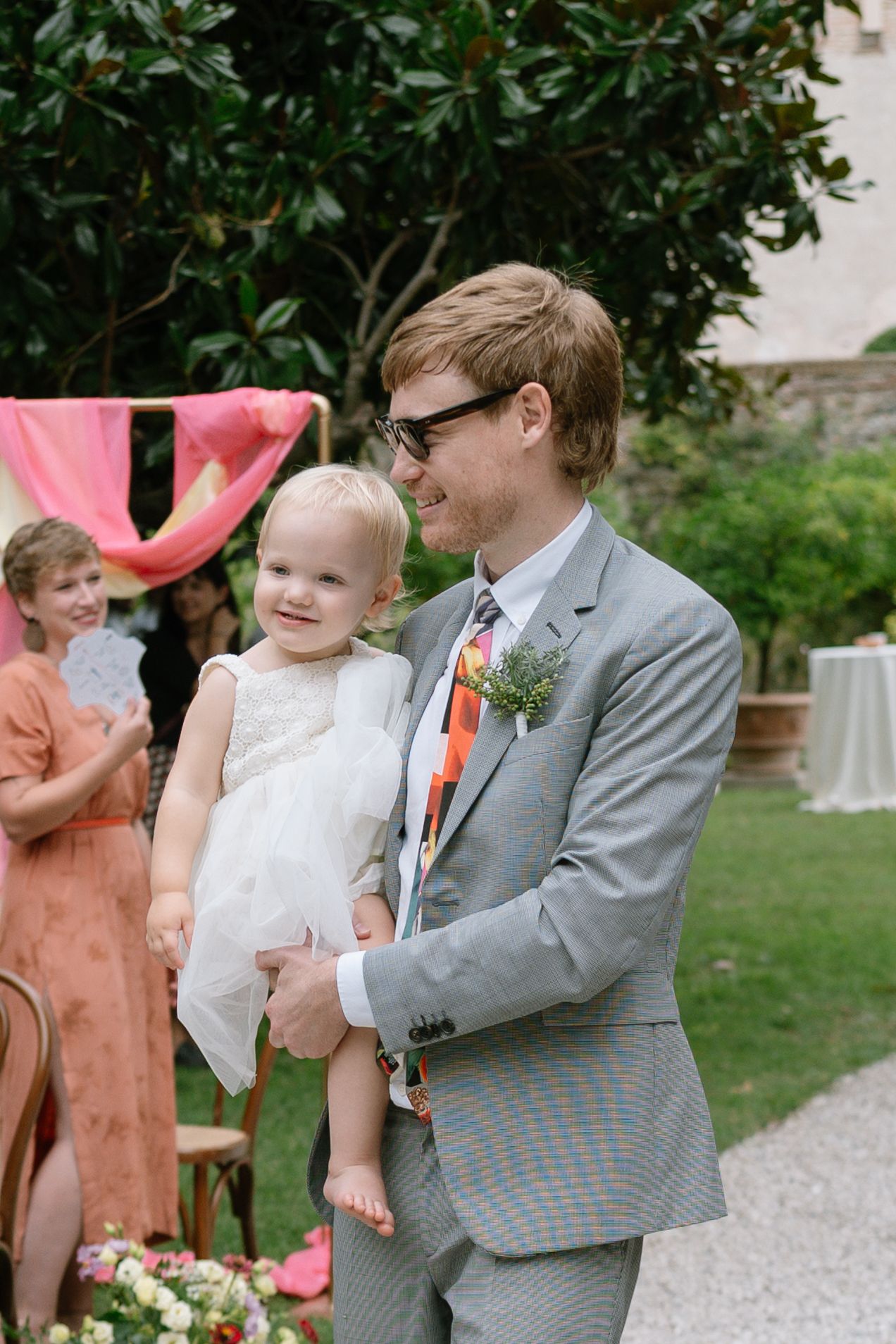 A couple shares a joyful moment during their outdoor wedding ceremony with guests in coral dresses looking on.