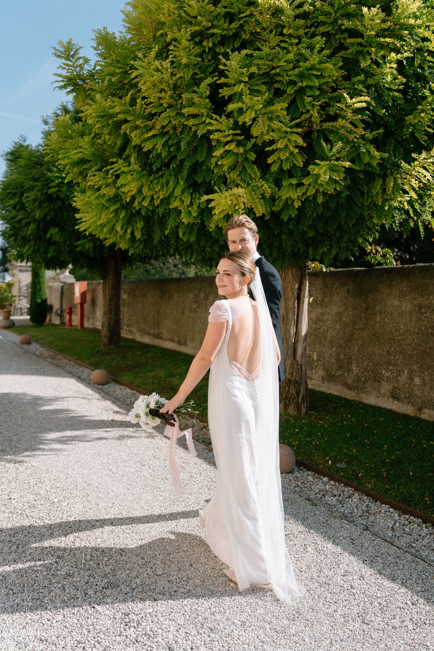 Bride in backless dress and groom stroll along scenic tree-lined path.