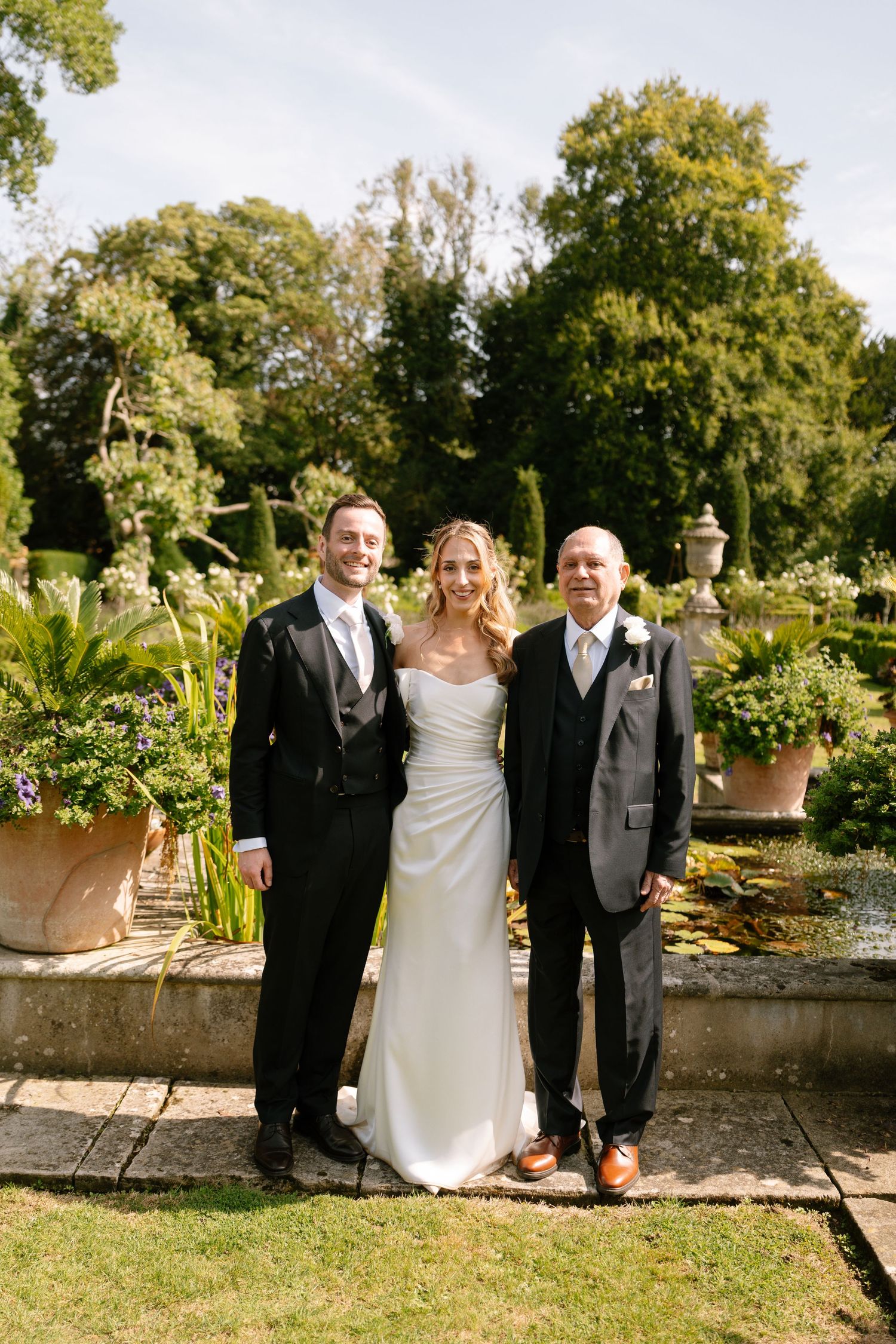 Wedding party poses for formal photos in a sunny garden setting.
