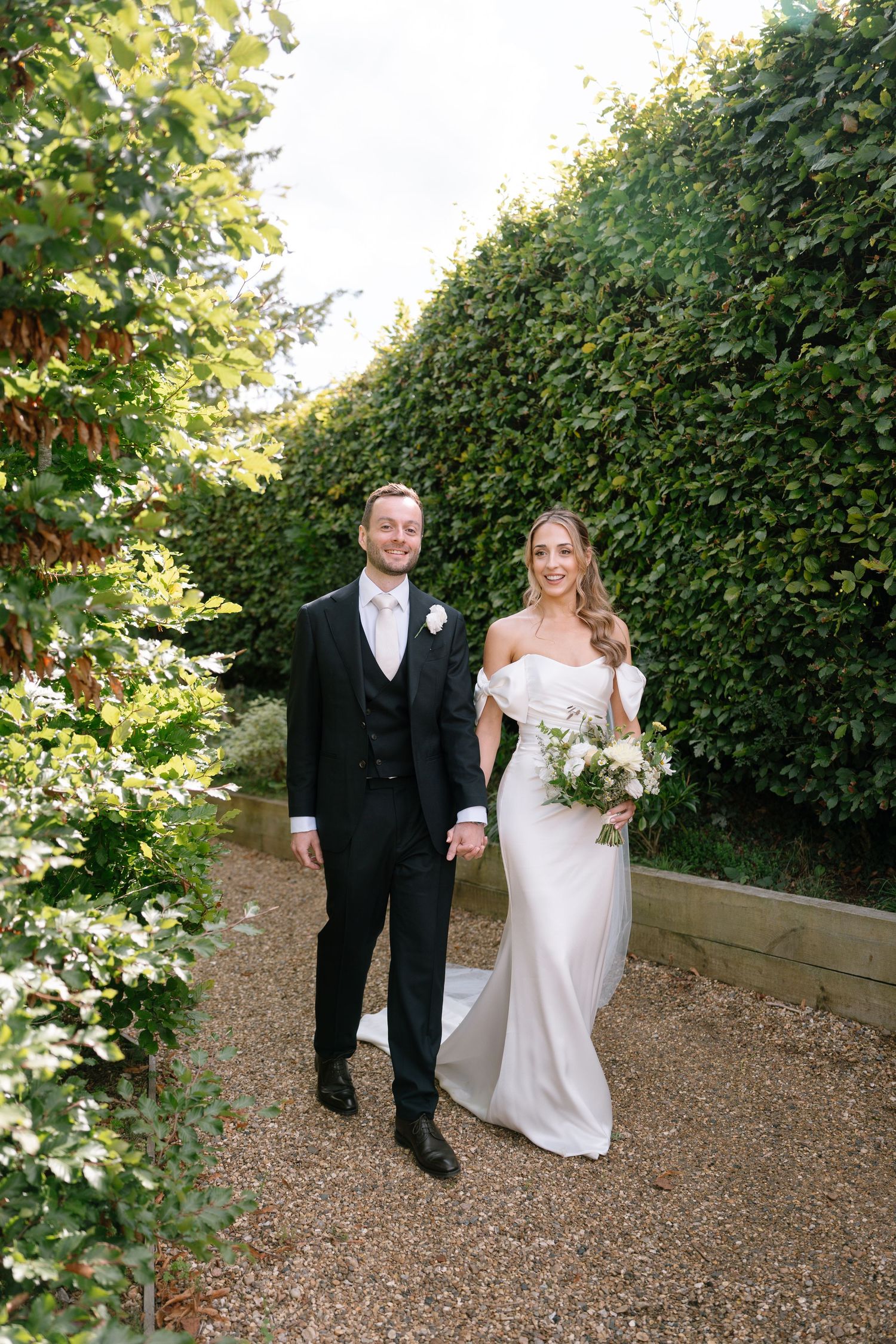 Wedding couple strolling down garden pathway with manicured hedges and gravel ground.