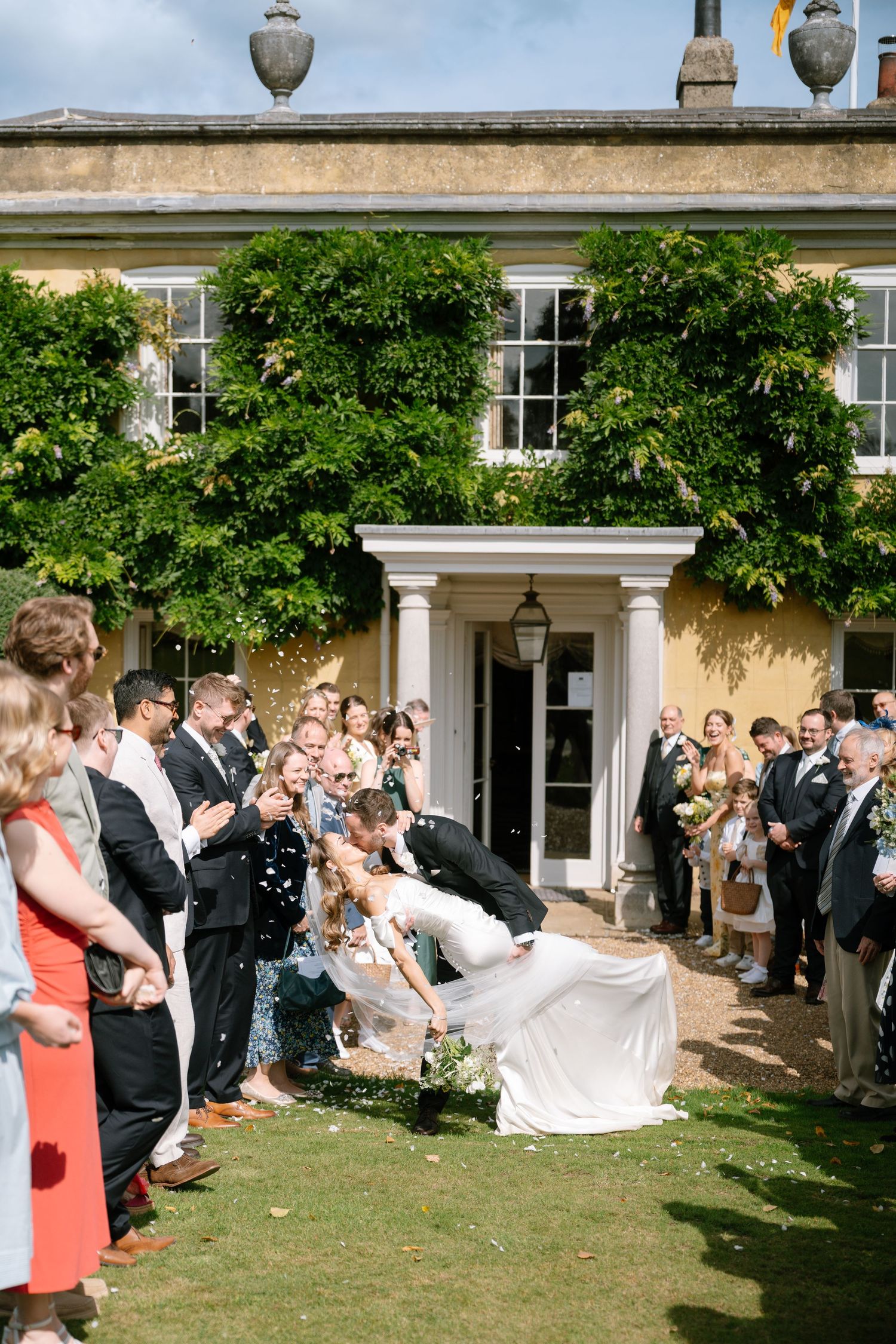 Wedding party and guests celebrate with confetti on the lawn of an elegant venue.