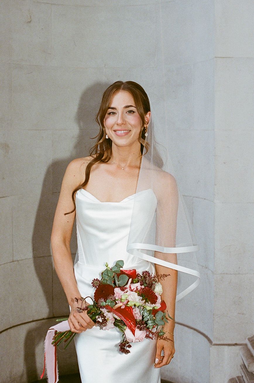 Smiling bride in strapless white gown holding red and pink floral bouquet with ribbon, standing against stone wall.